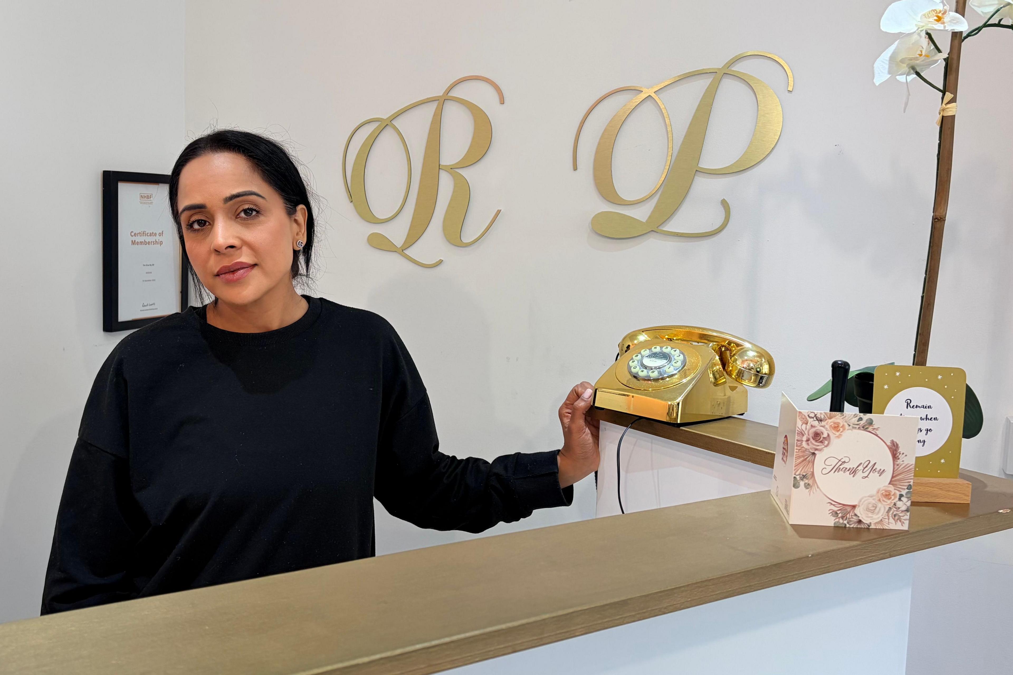 A woman in a black sweater stands behind a counter and looks at the camera. A gold telephone, two cards and a white plant are on the desk. Two gold letters - 'r' and 'p' are hung up on the wall alongside a certificate.