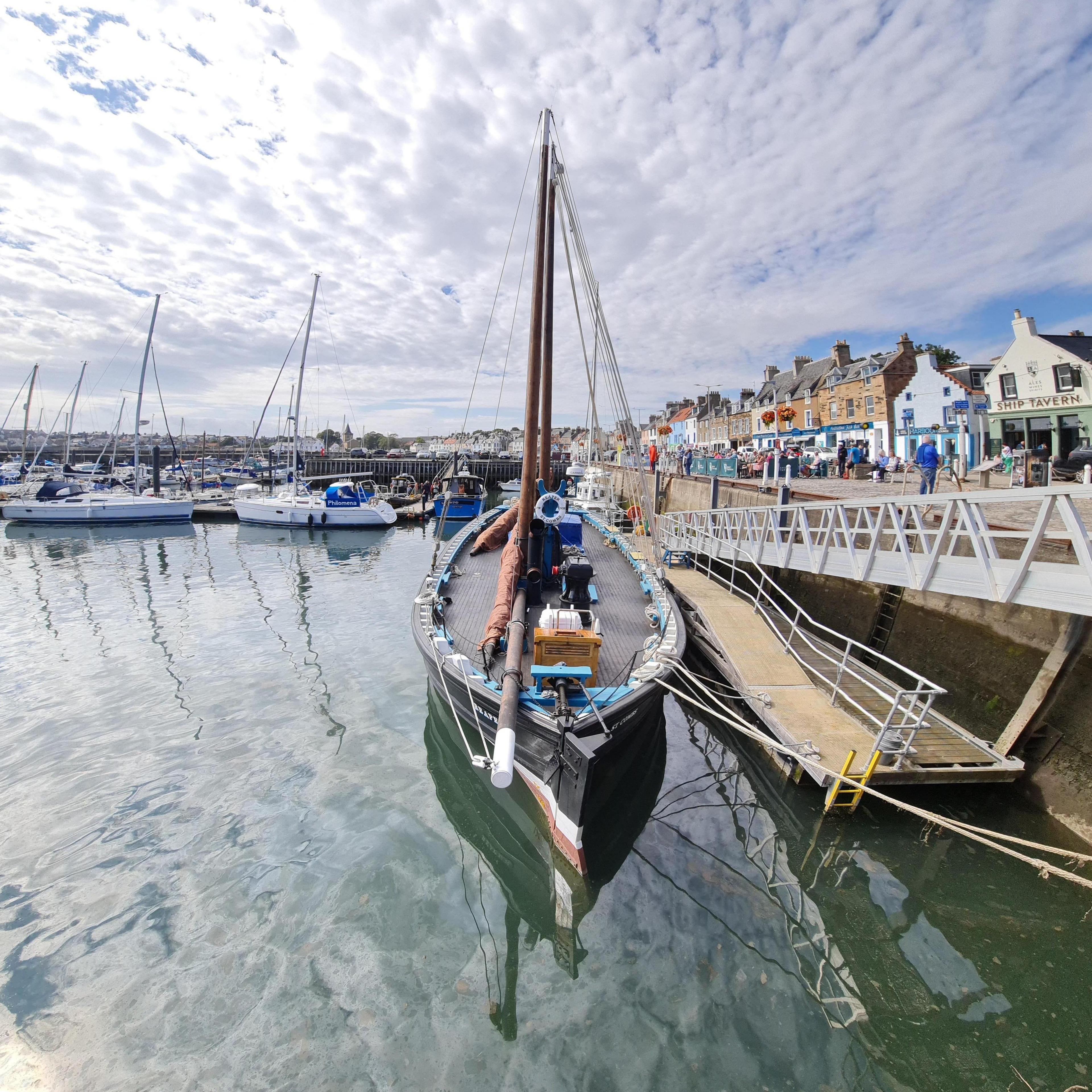 A boat with a black hull sitting on calm, reflective water which has reflected the cloudy sky. The boat has two large masts on the deck next to a light-coloured gangway. In the background are a number of other boats and some colourful buildings.