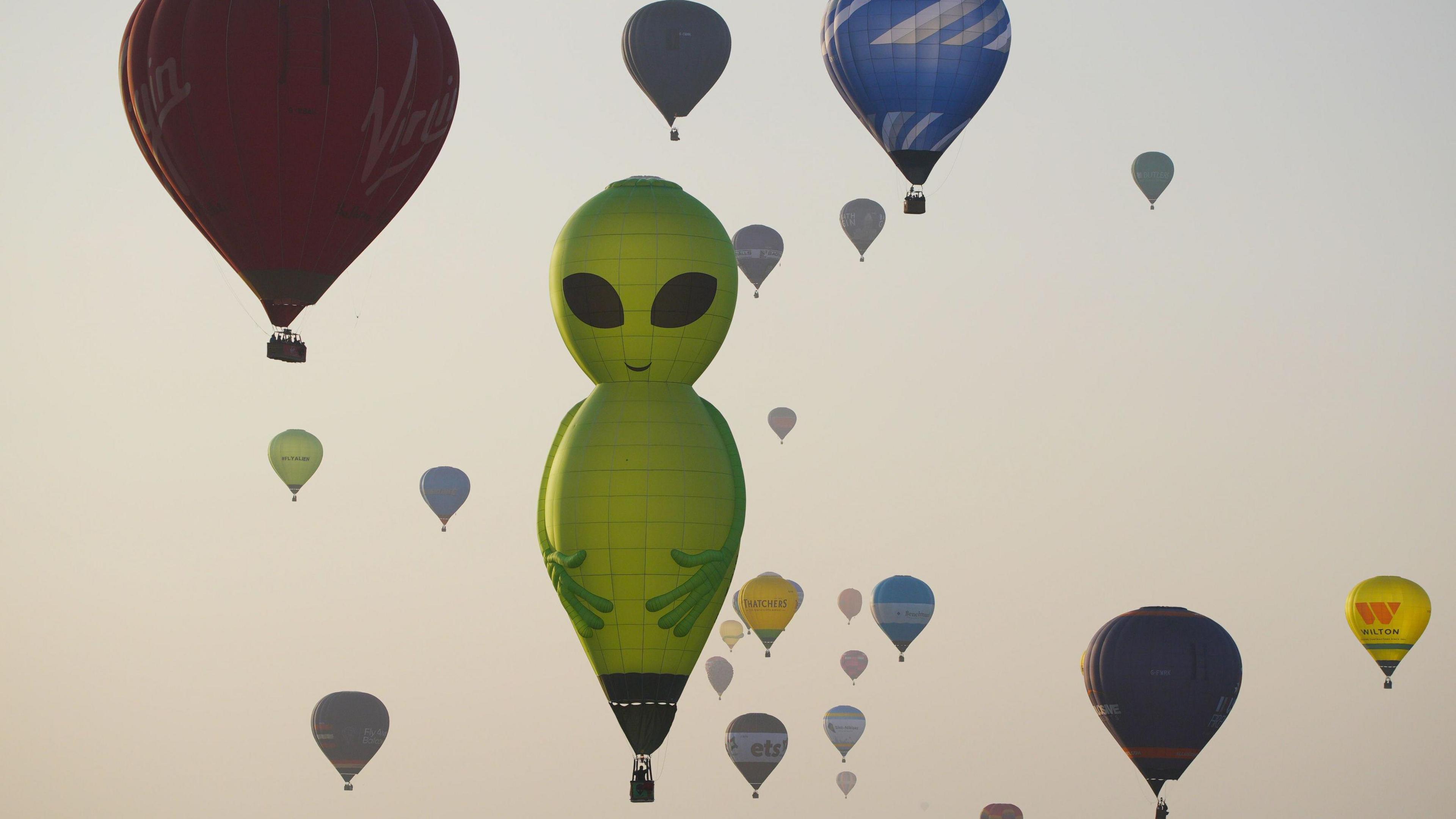 Dozens of balloons of all shapes and colours rise into the pale, clear morning sky at sunrise. The main focal point is the large, green balloon in the shape of an alien with black eyes.