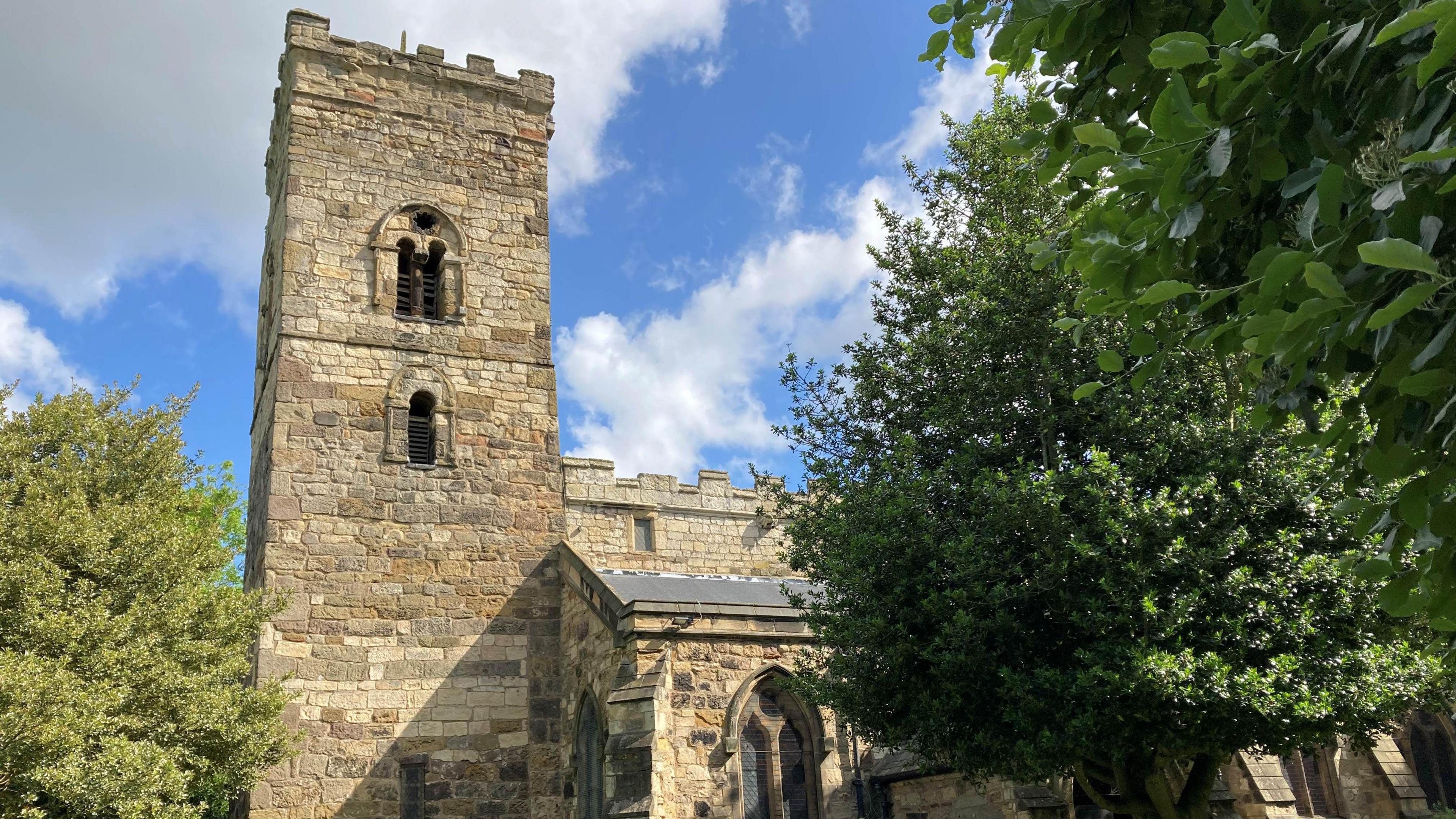The crenelated tower of St Cuthbert's Church emerging over a tree. The roof of the main body of the church can also be seen.