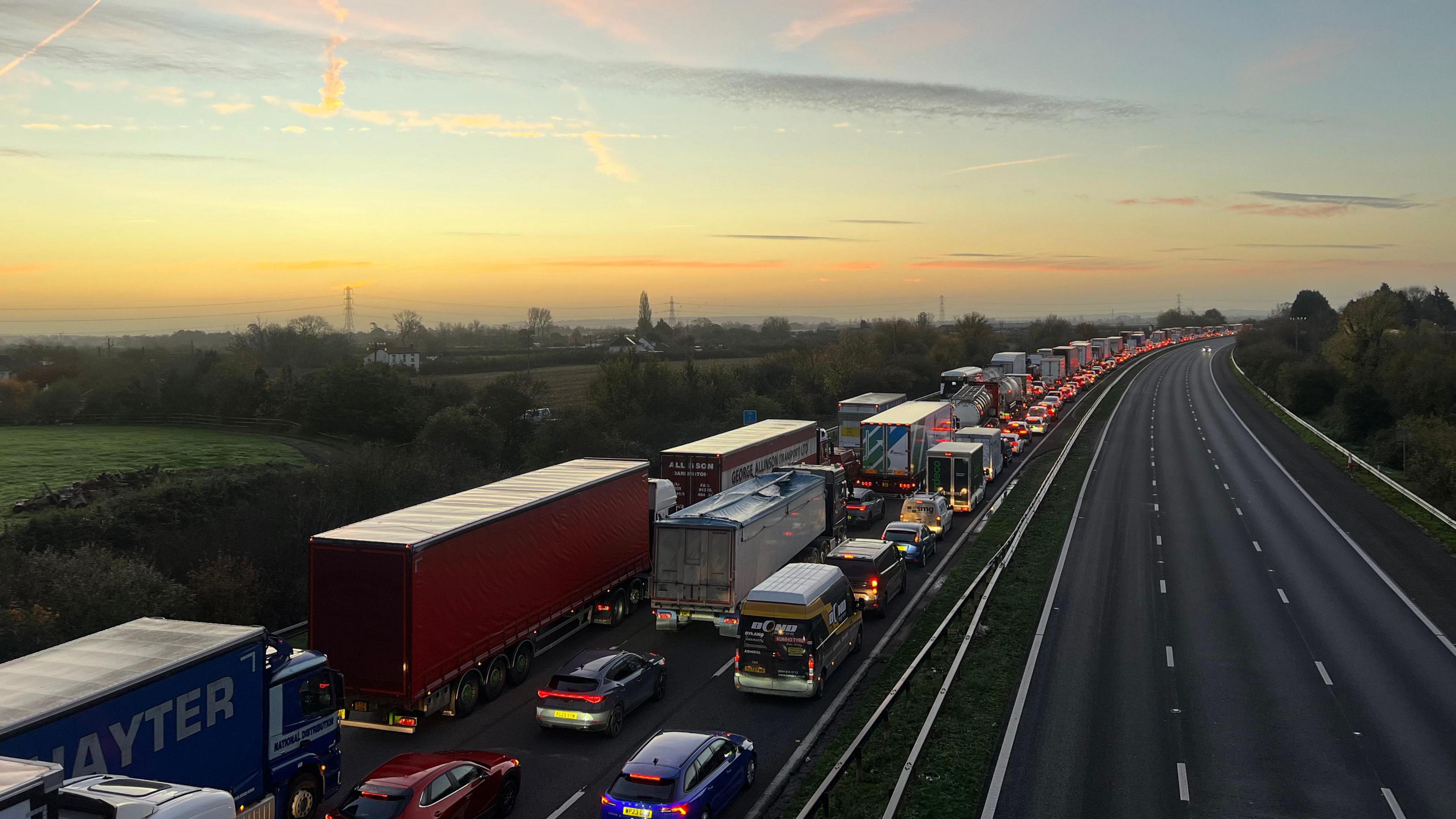 Lines of traffic on the M5 motorway. The road is blocked full of vehicles, which can be seen for miles. The sun is rising in the background.