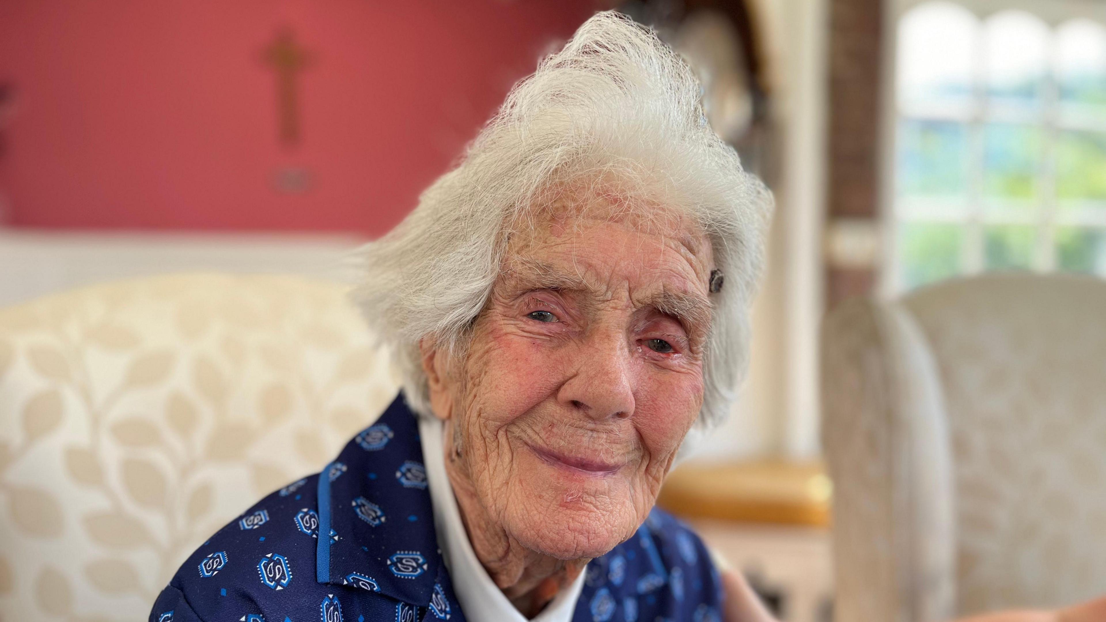 Joan Willett looks at the camera smiling. She has white hair and is wearing a patterned blue jacket over a white shirt.