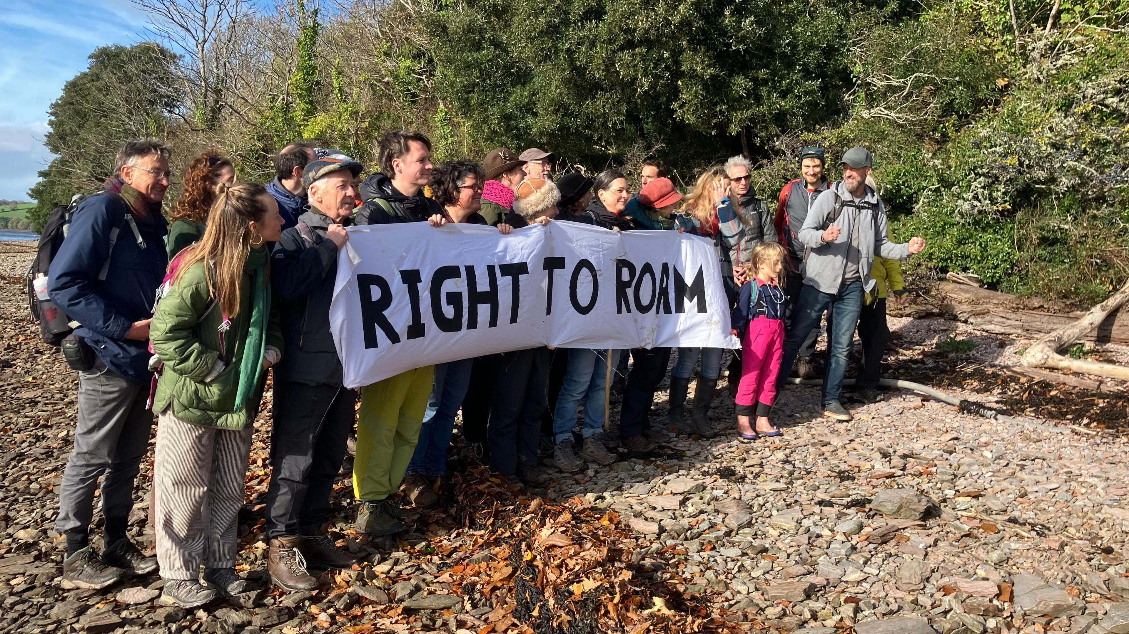 A group of people stood together holding a banner which says RIGHT TO ROAM. They are standing on the banks of a river and are wearing backpacks and coats.