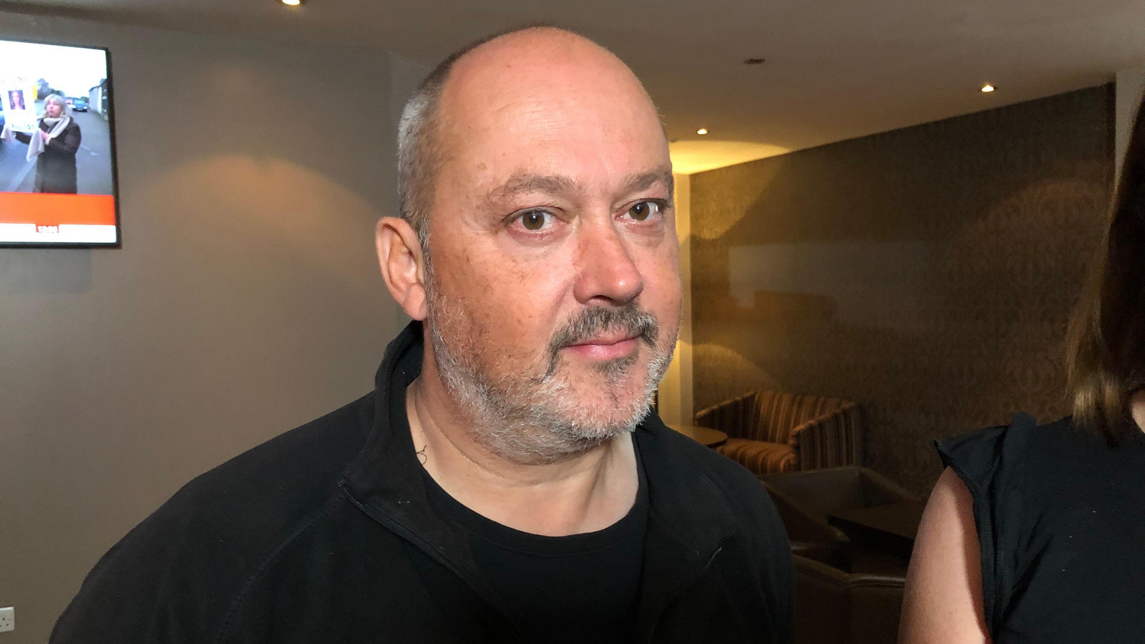 Stuart Stephens, a bald man with a grey beard, wearing a black top and looking directly at the camera.