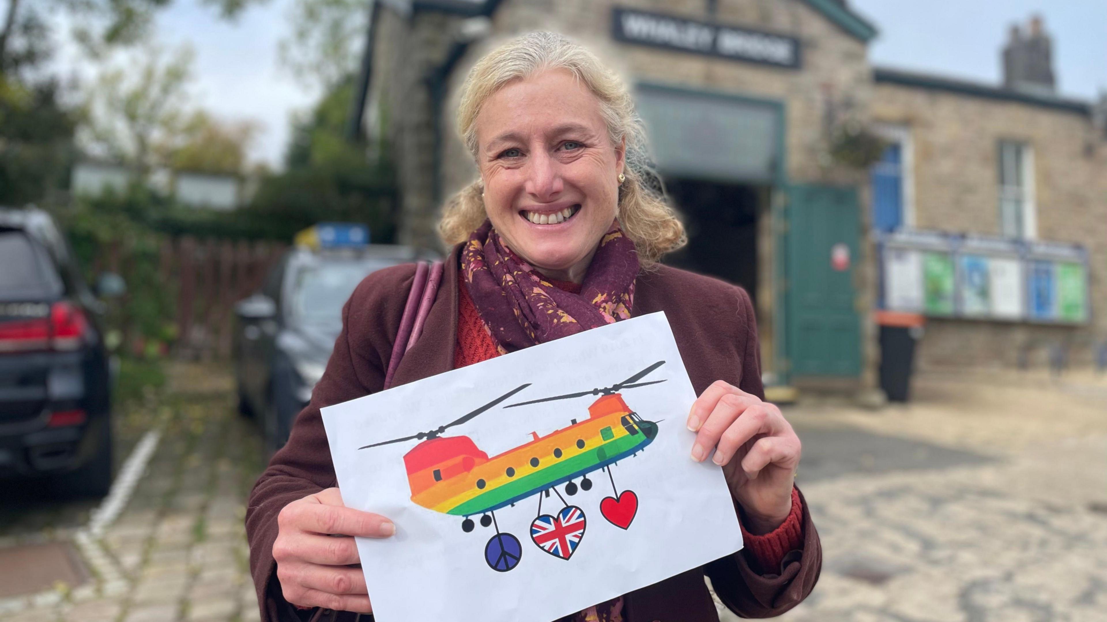 Woman in a claret jacket holding a picture of a multi-coloured Chinook helicopter