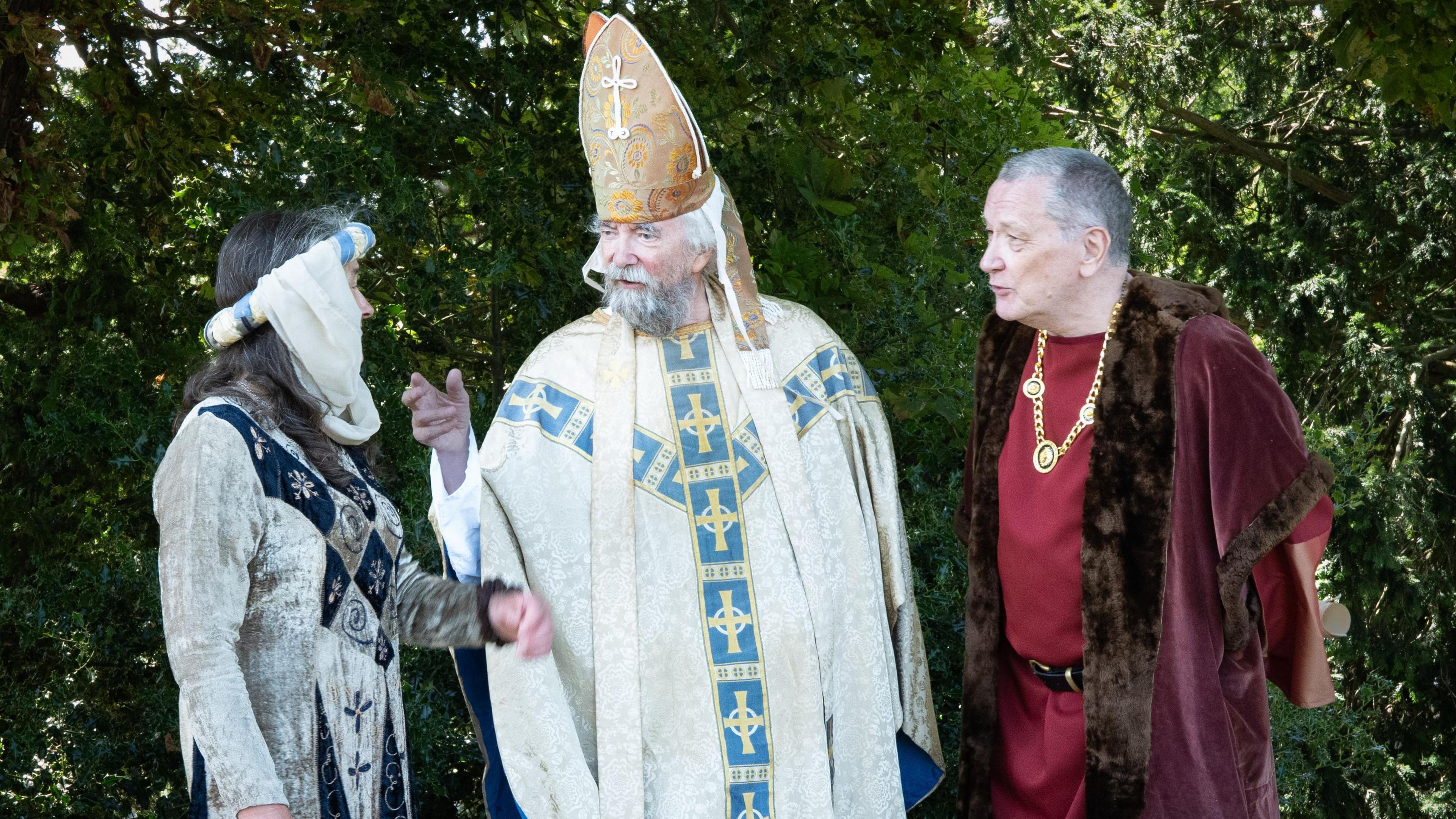 Three characters in costume. A lady is wearing a medieval dress with a headdress, a man is in church attire, and another man is wearing a burgundy costume with fur trim and a chain