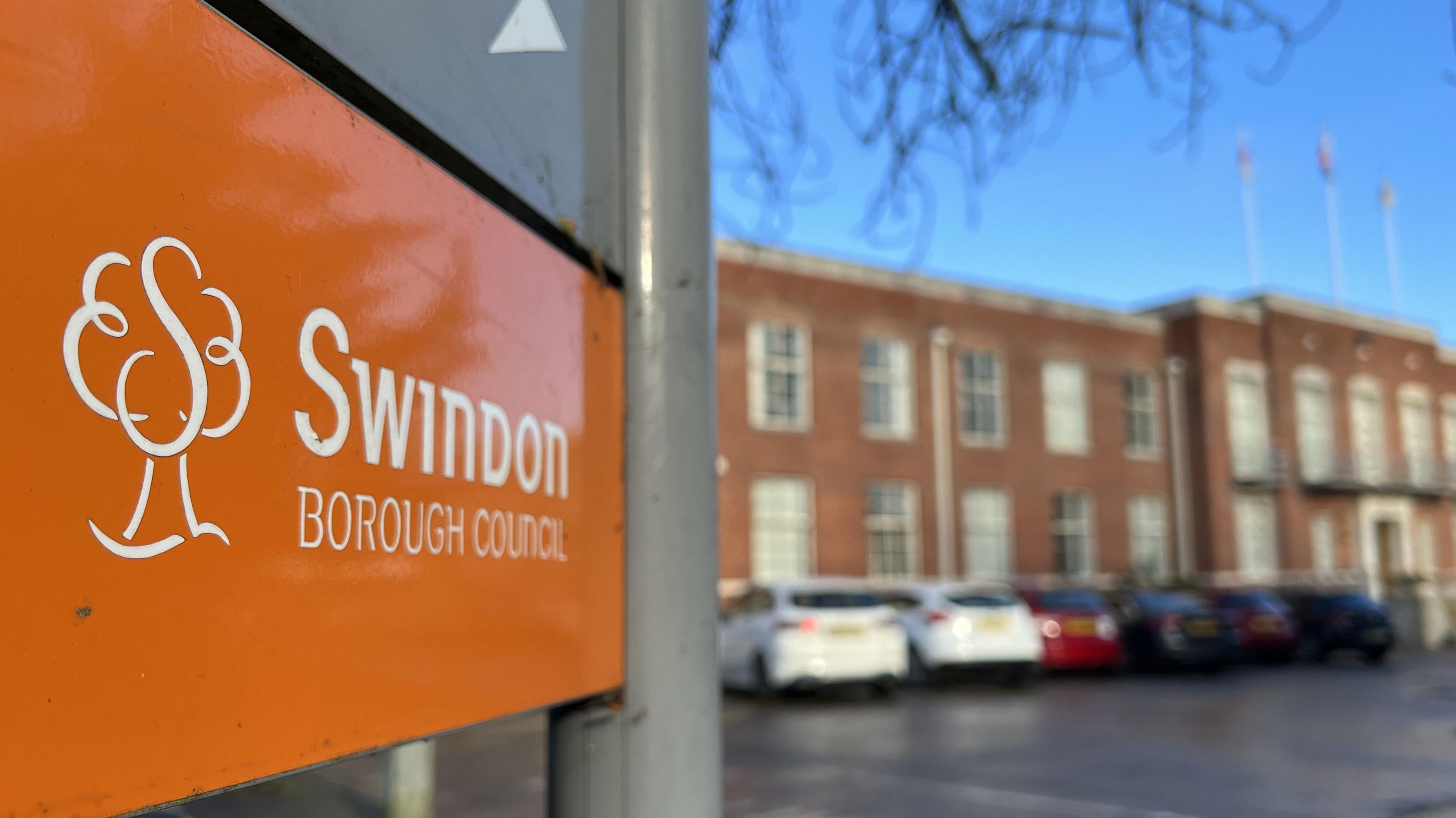 An orange Swindon Borough Council sign outside the brown brick council offices. The sign is in focus, while the building behind is blurred, with cars parked in front of it.