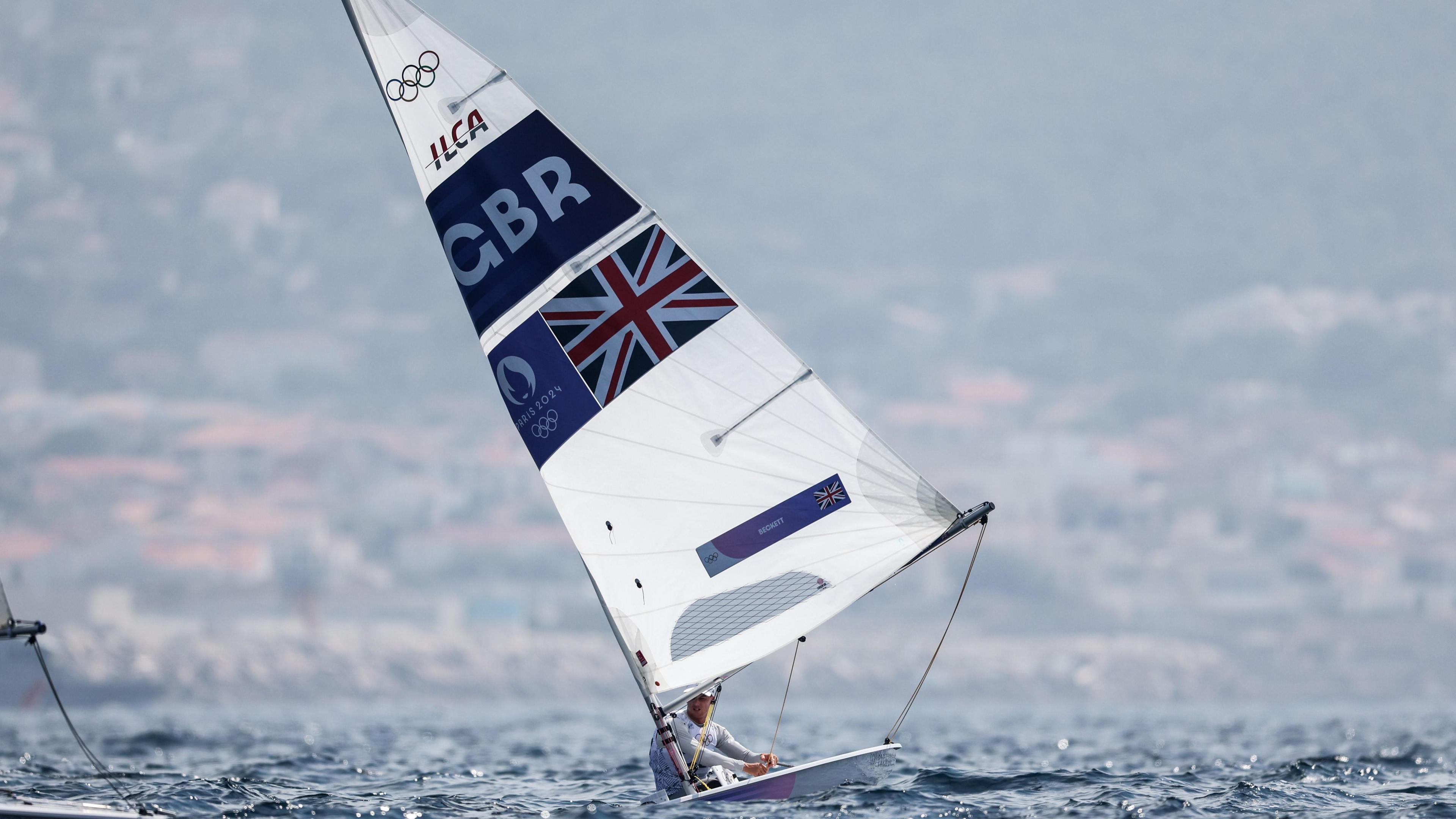 Mickey Beckett on his boat during the Paris 2024 olympics. It is small, with a large sail with the union jack and GBR on it.