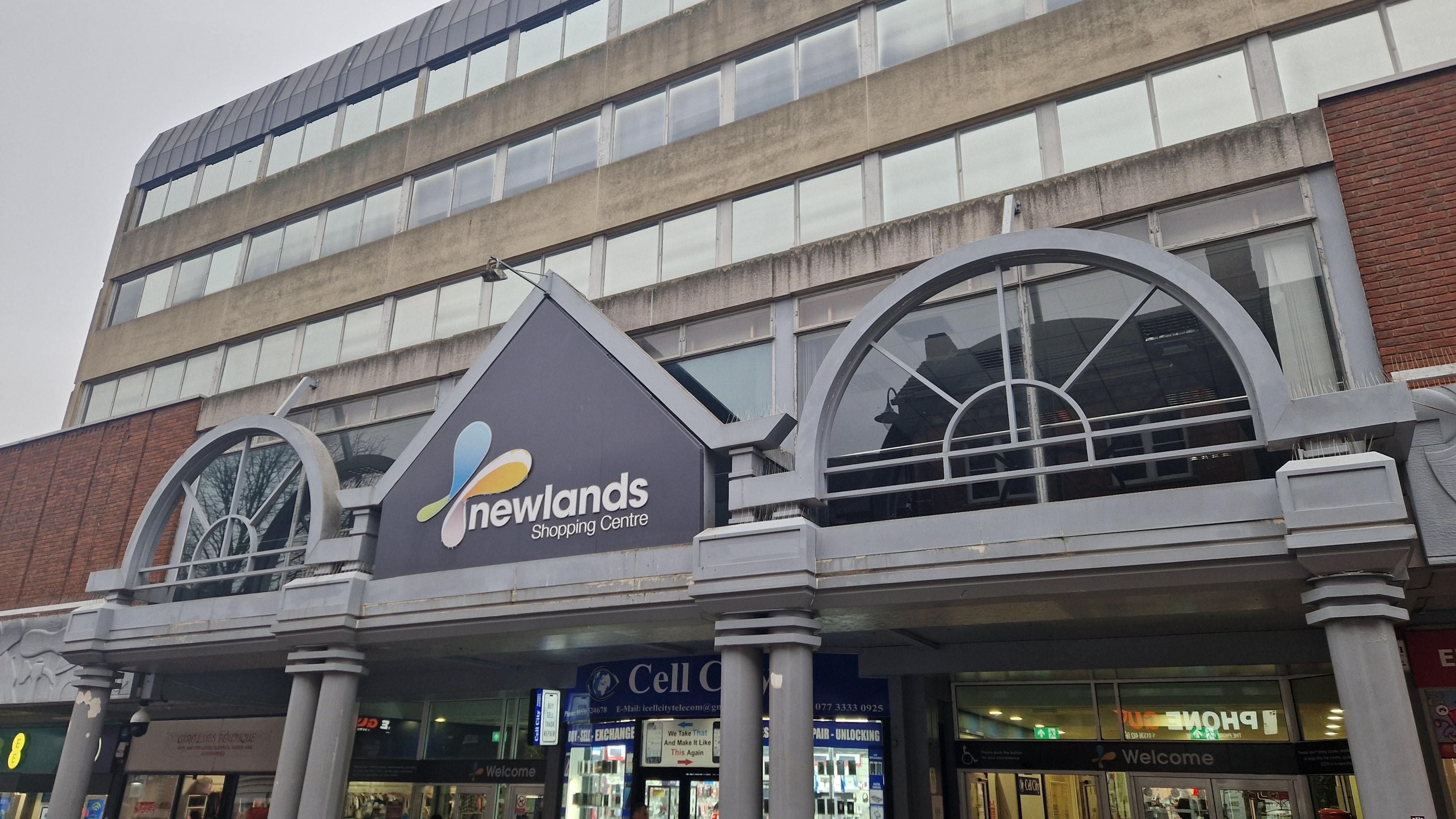 Newlands Shopping Centre, showing a sign and an entrance to the centre, including modern arch ways, shops and a brick building with several floors.