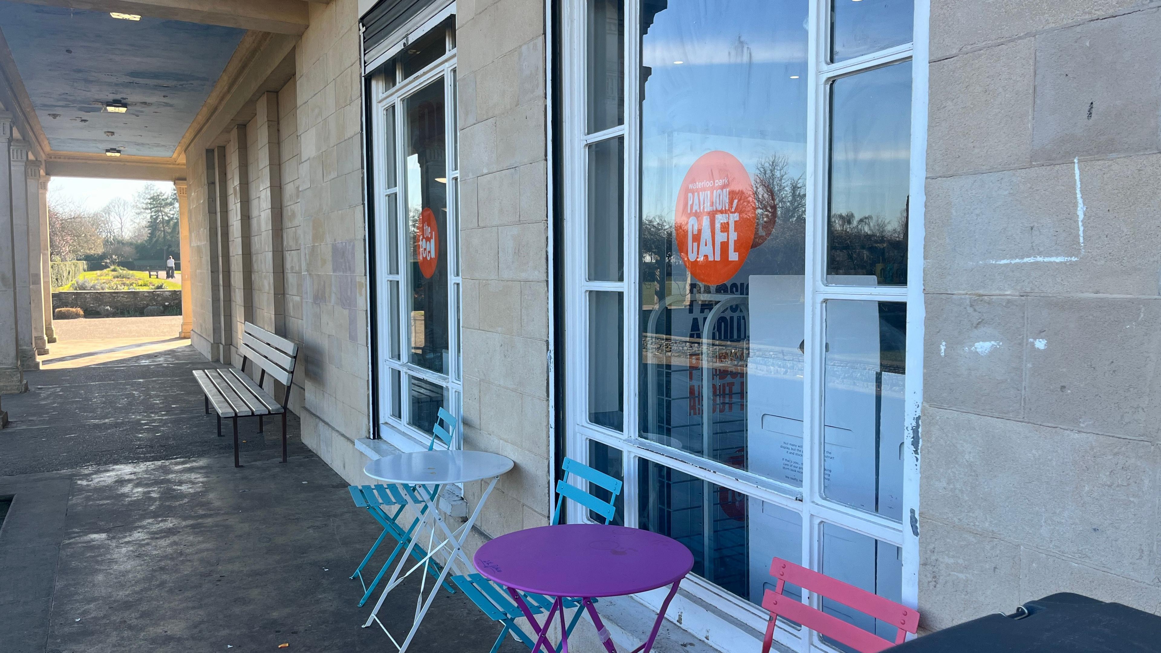 The front entrance of The Feed pavilion cafe. Outside is a bench, blue, white and pink metal chairs, and a purple table. On the windows are orange signs with the cafe's branding on them.