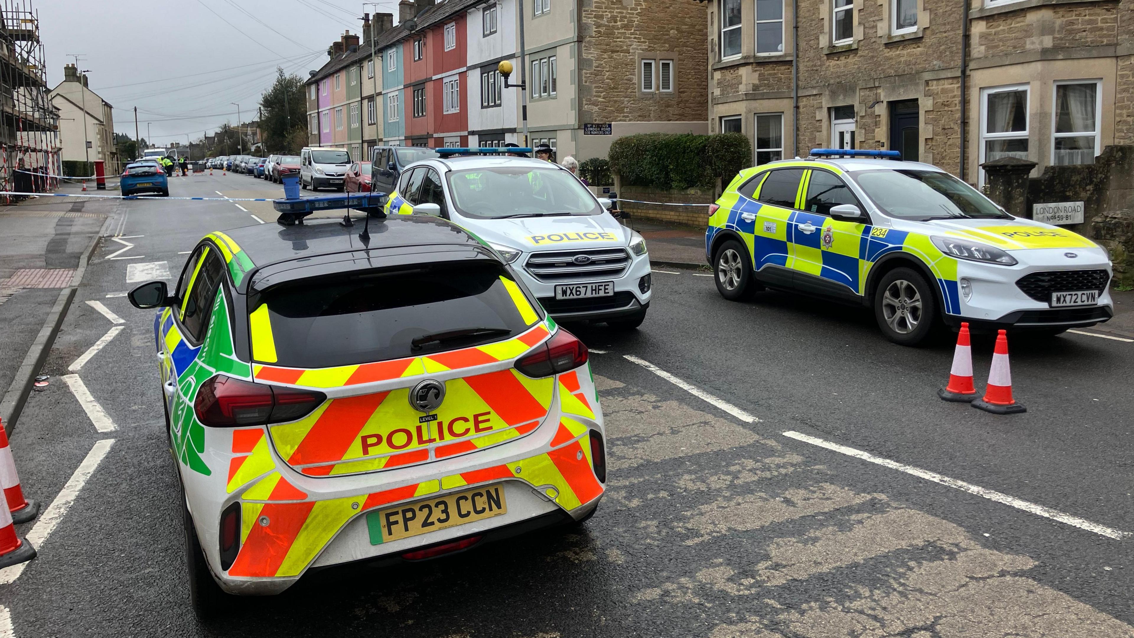 Three marked police cars block a residential street. There is blue and white police tape across the road. It is a grey day, and some of the terraced houses are painted bright pastel colours. 