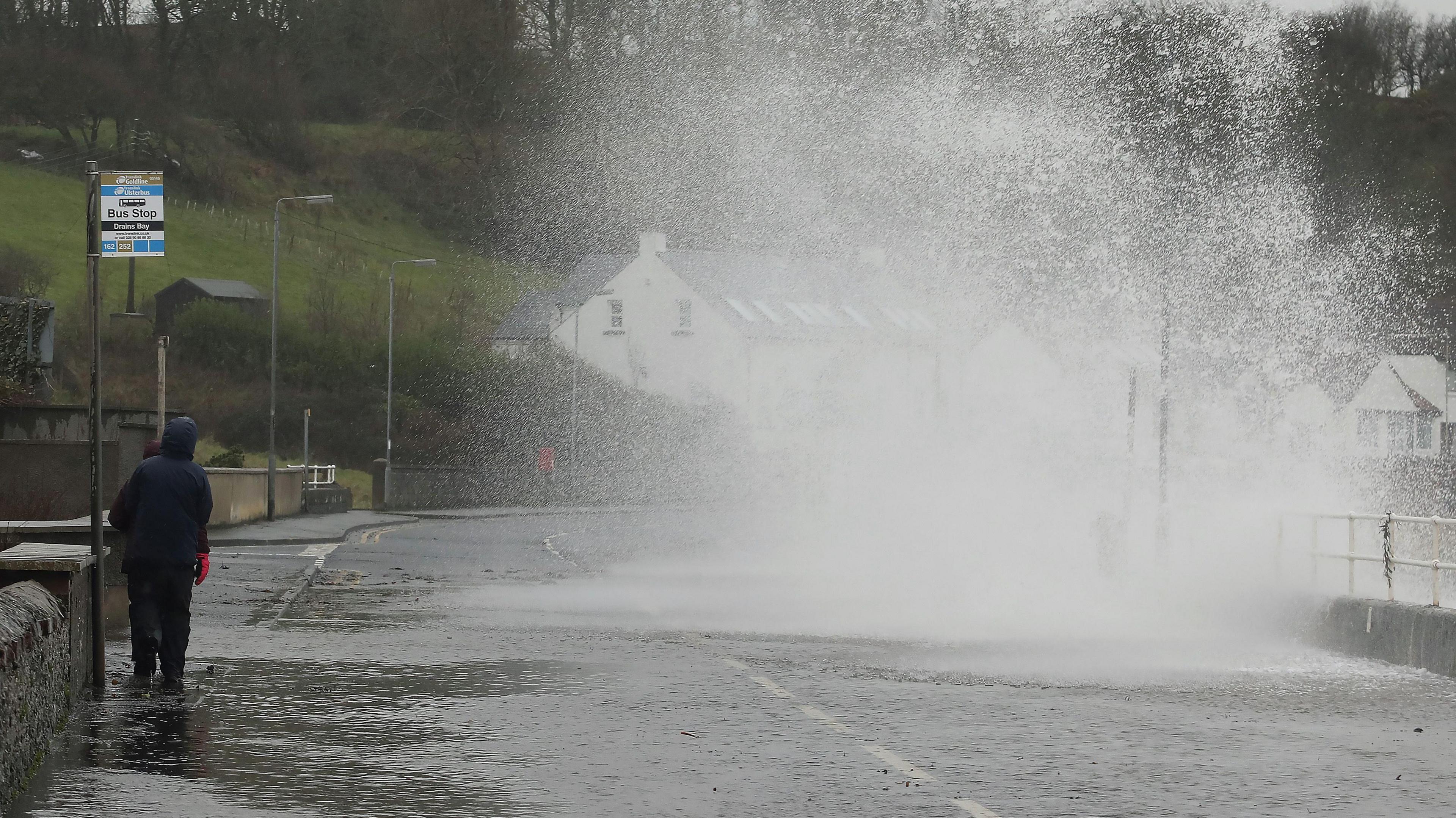 Waves crash onto a coastal road. A man is walking passed a bus stop. 