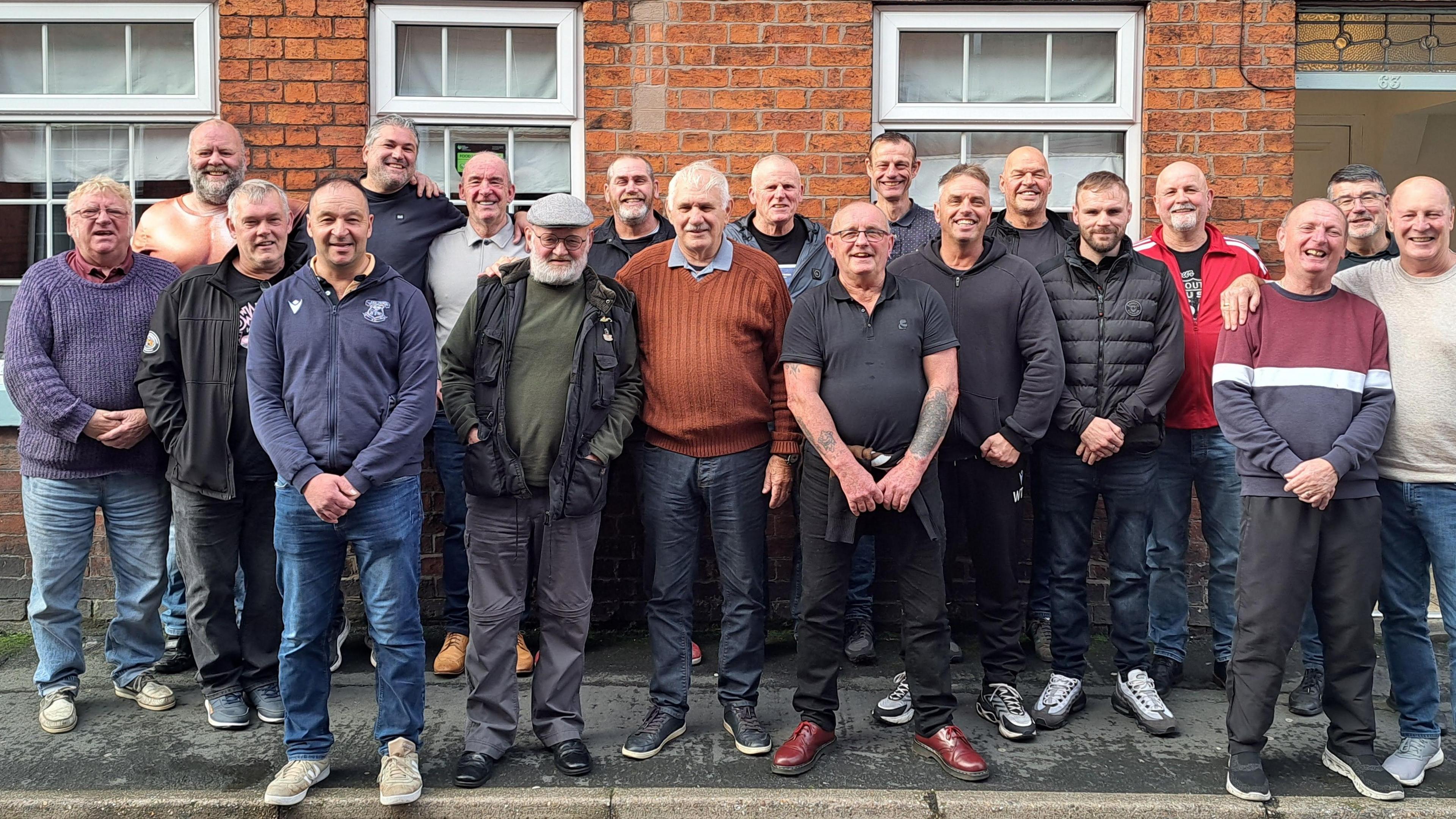 Twenty men stand on the pavement, gathering in a huddle outside a brick building. Three window panes and a door can be seen behind them. They are all dressed differently, mainly in jackets, dark jeans and trainers.