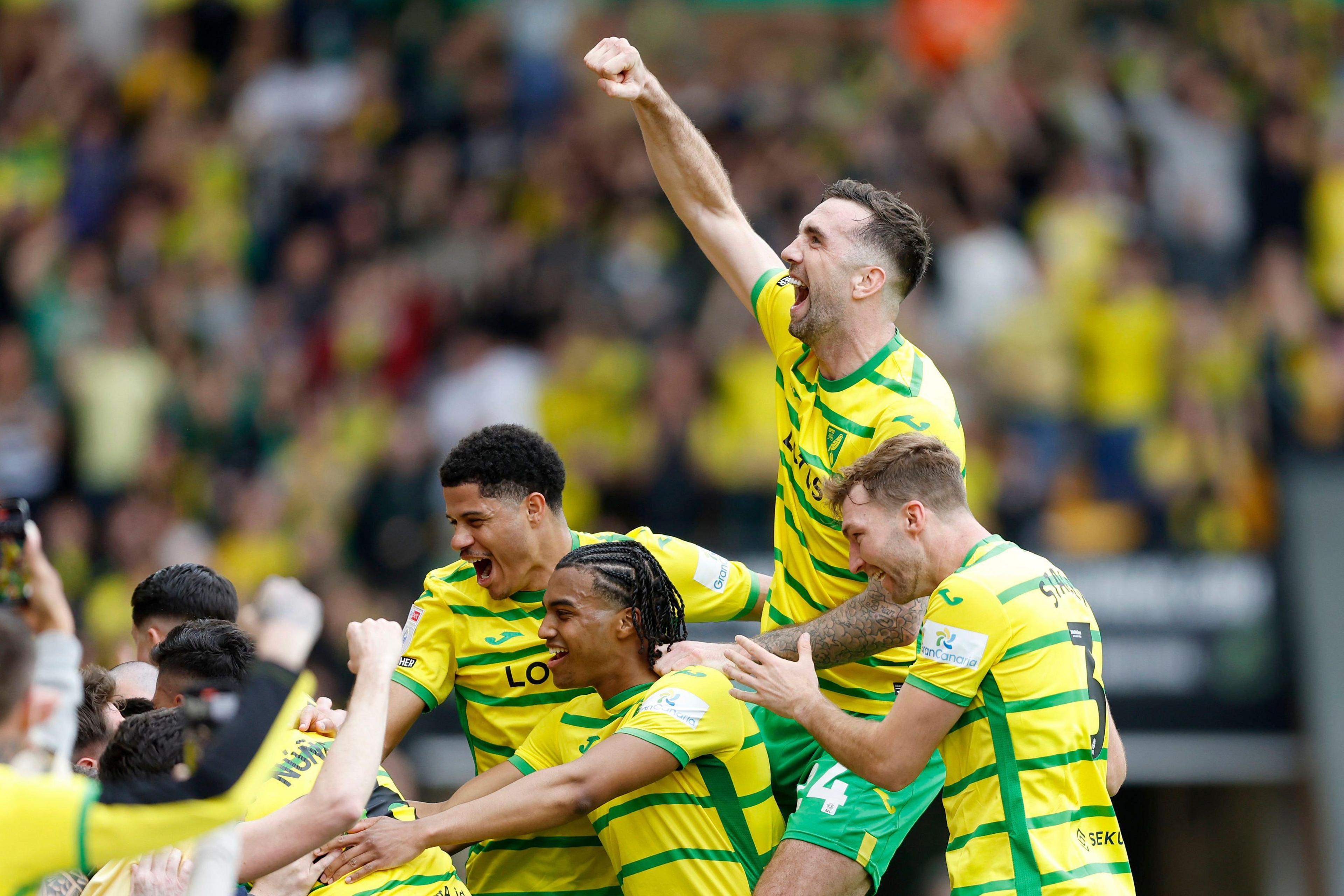 Four football players celebrate a goal as their team-mates hug supporters at the front of a stadium's stand.