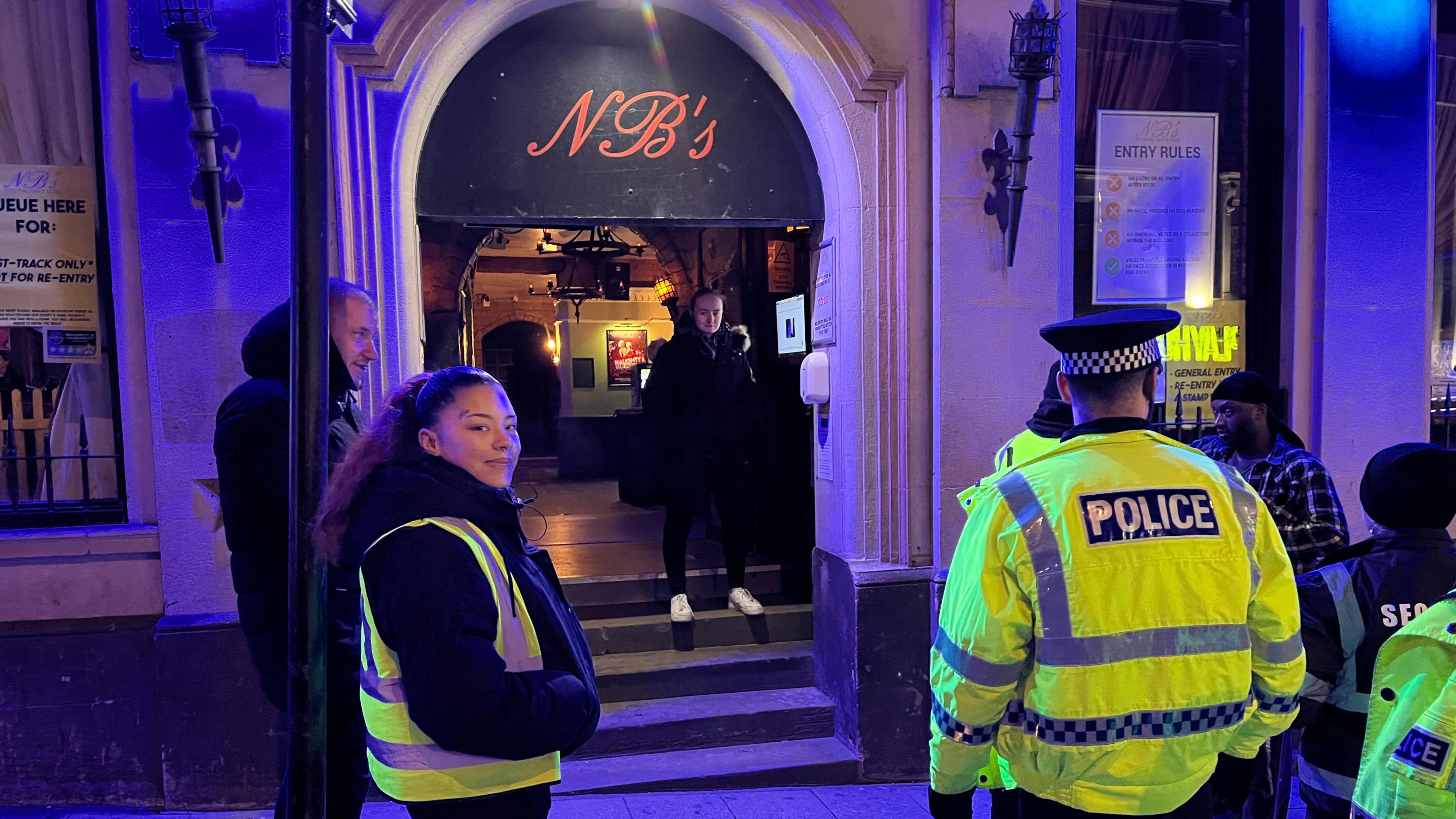 Two police officers in green high visibility jackets and a black police hat look at the entrance of a night club. A man stands on the first step to the club's entrance. A member of the doorstaff looks at the camera.