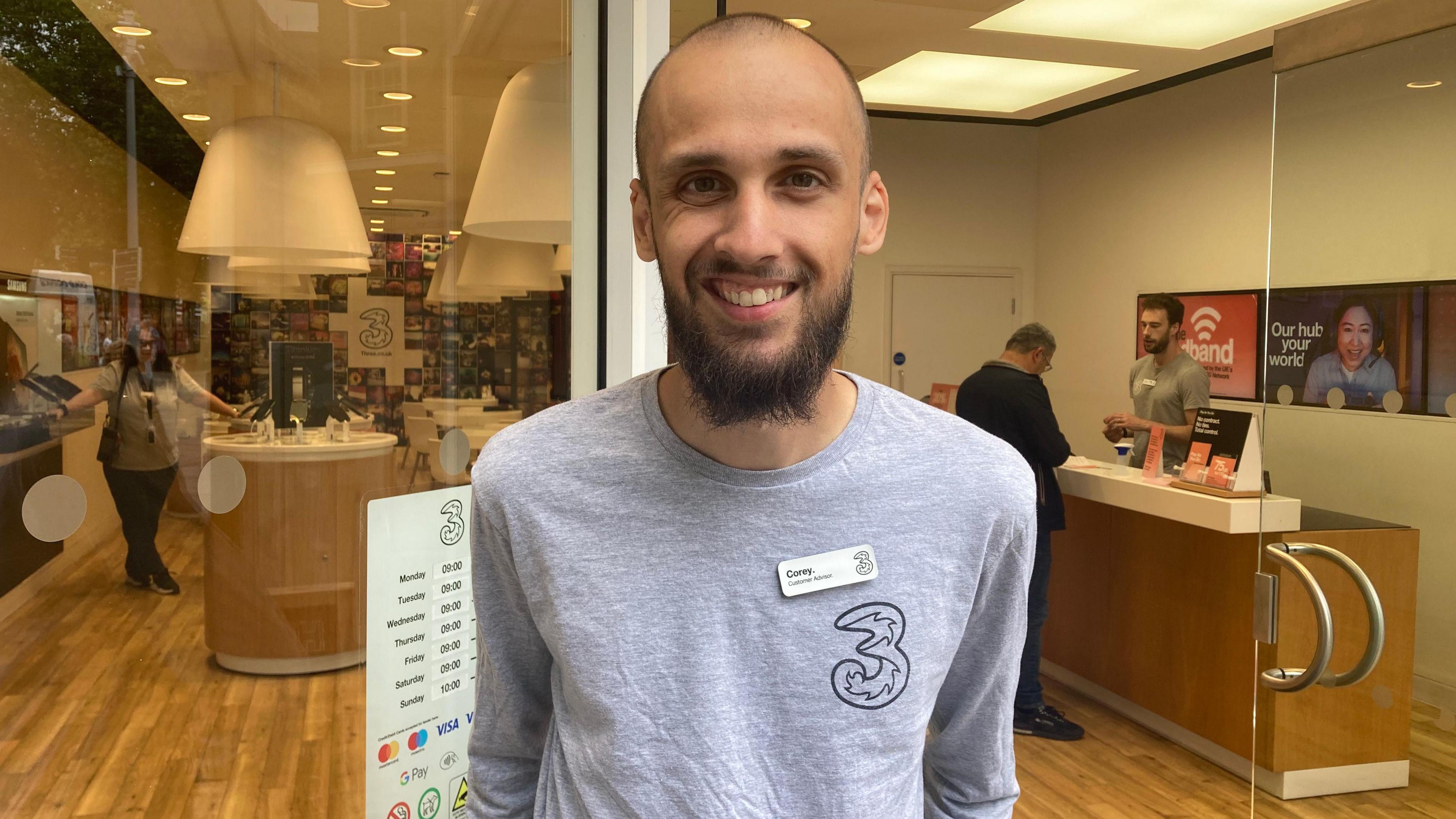 Corey Hunter-Mitchell stands outside the Three shop in Hull. He is wearing a grey T-shirt and has a name badge. The store behind him has customers looking at phones and talking to staff