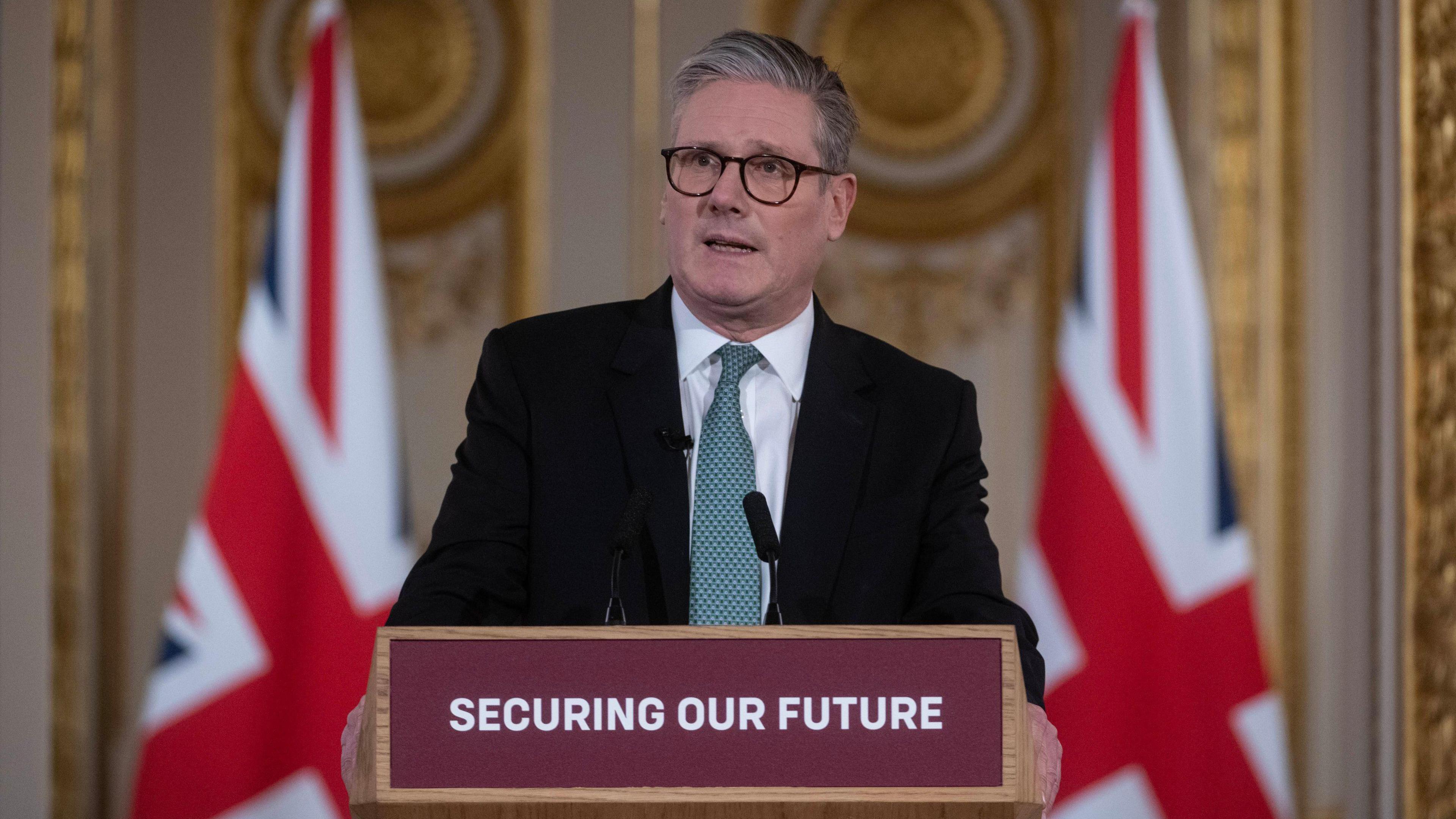 Sir Keir Starmer wearing a black suit and white shirt with green tie, standing in front of two Union Jack flags and behind a podium with the slogan "securing our future".