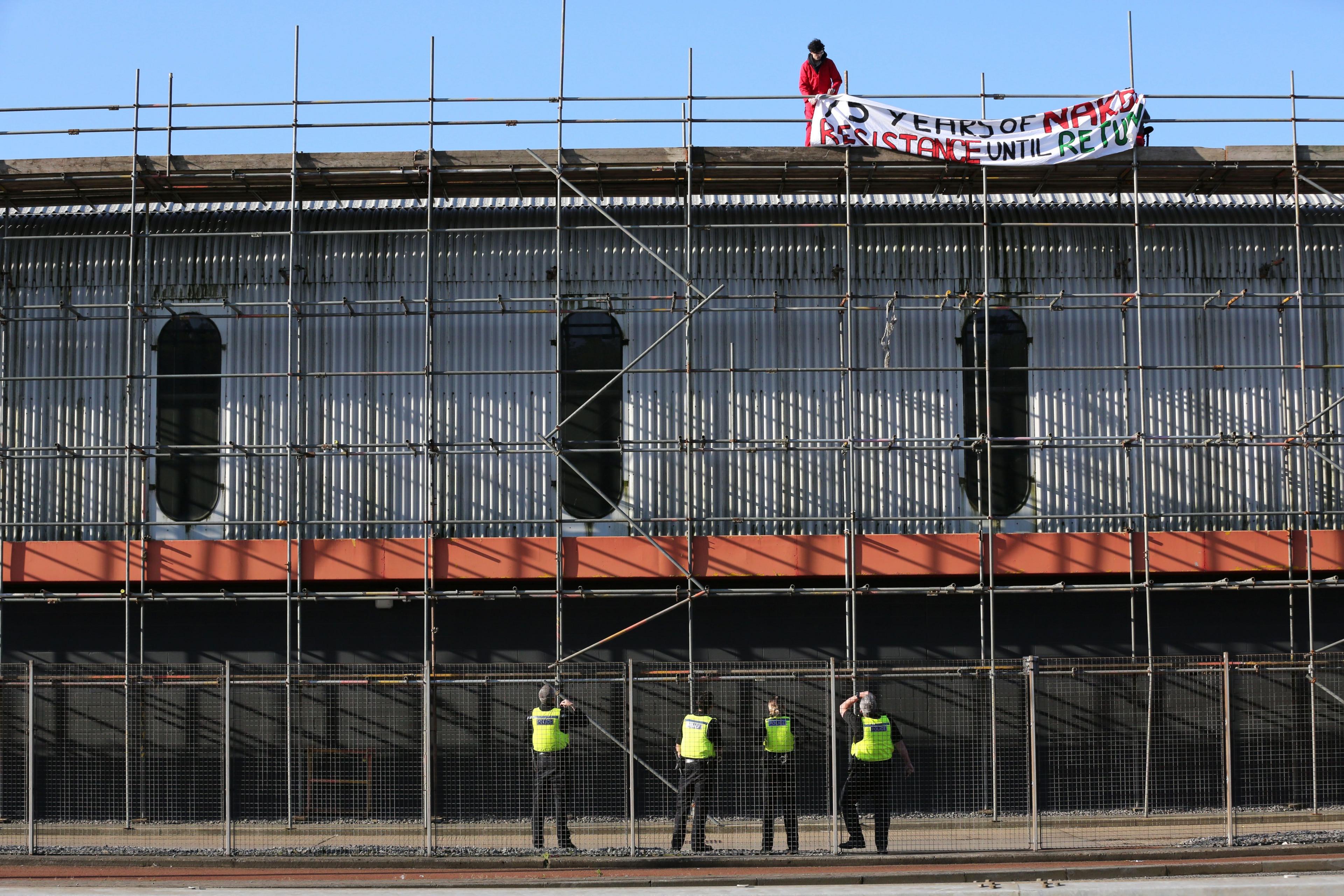 Four police officers on the ground watch as a man unfurls a banner on a rooftop several stories high