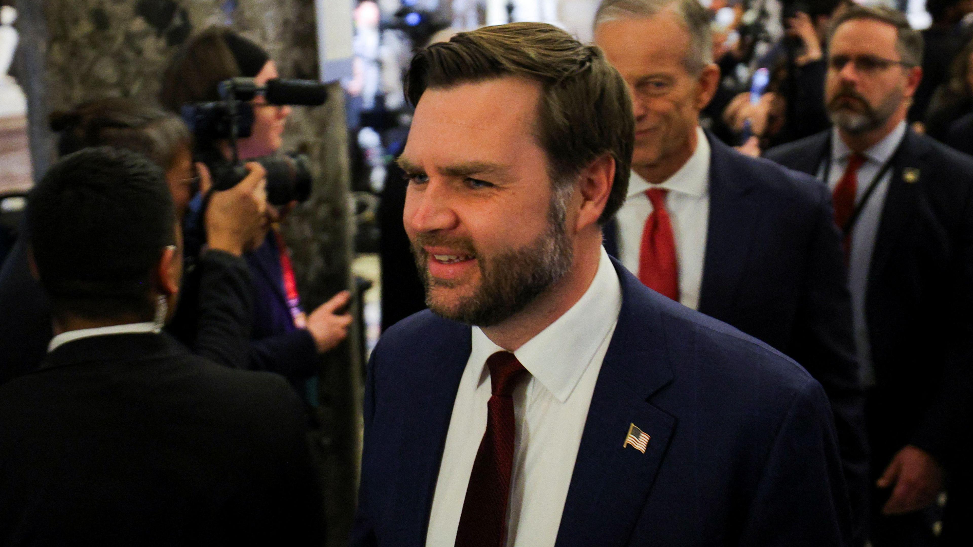 Vice president JD Vance, who has short brown hair and a short beard and moustache, wearing a navy blazer, white shirt and red tie. He is walking through a crowd of reporters.