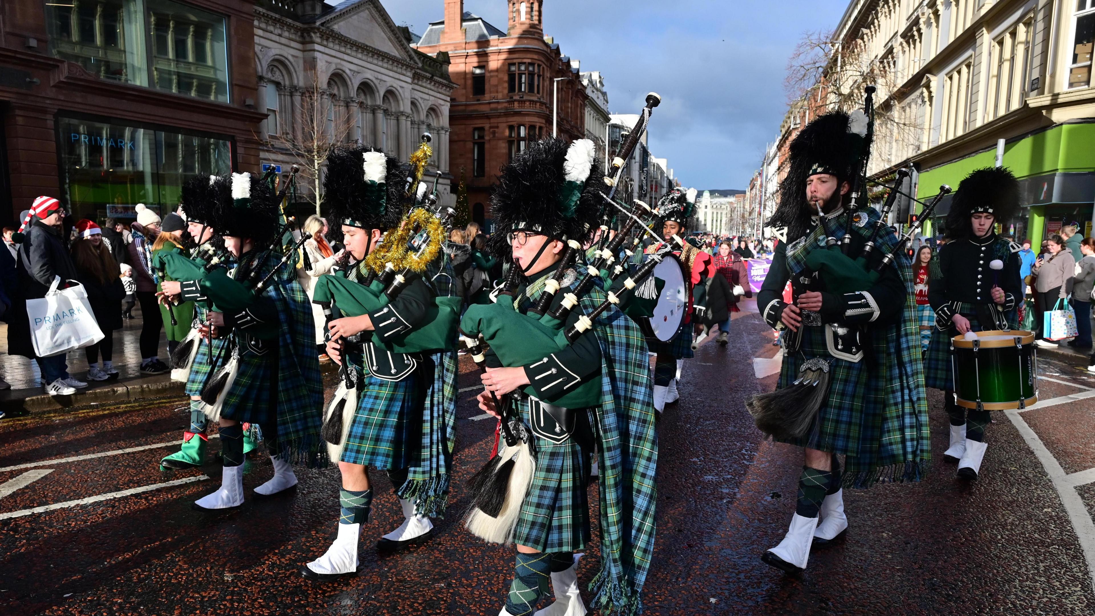 The picture shows a number of men playing bagpipes through Belfast city centre. They are wearing beefeater type hats and green tartan 