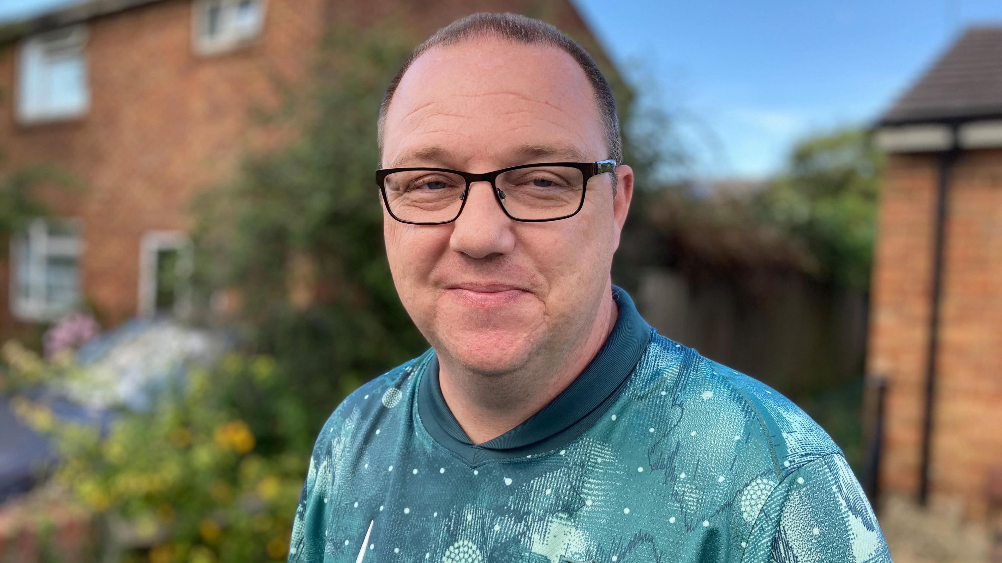A man wearing a blue shirt and glasses is smiling at the camera. A house is out of focus behind him.