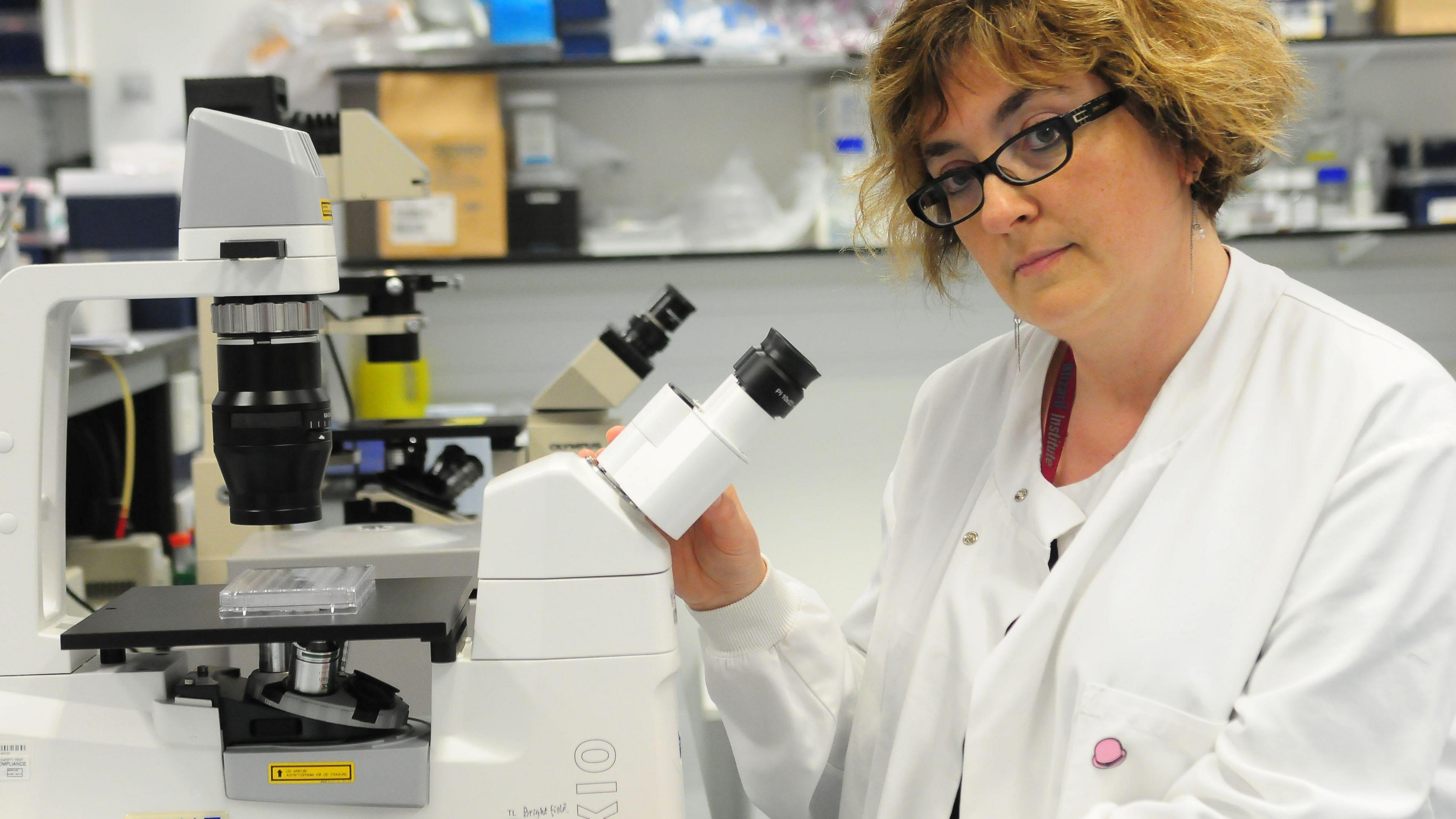 A woman in a lab coat and glasses stood next to a microscope. 