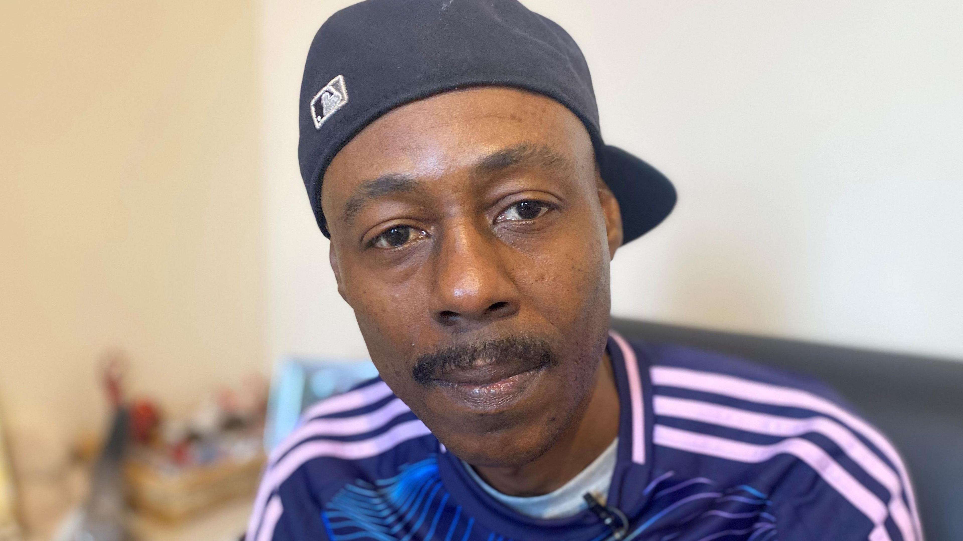 A man wearing a black baseball cap turned backwards wearing a blue jumper with white stripes along the shoulder and a white t-shirt underneath it