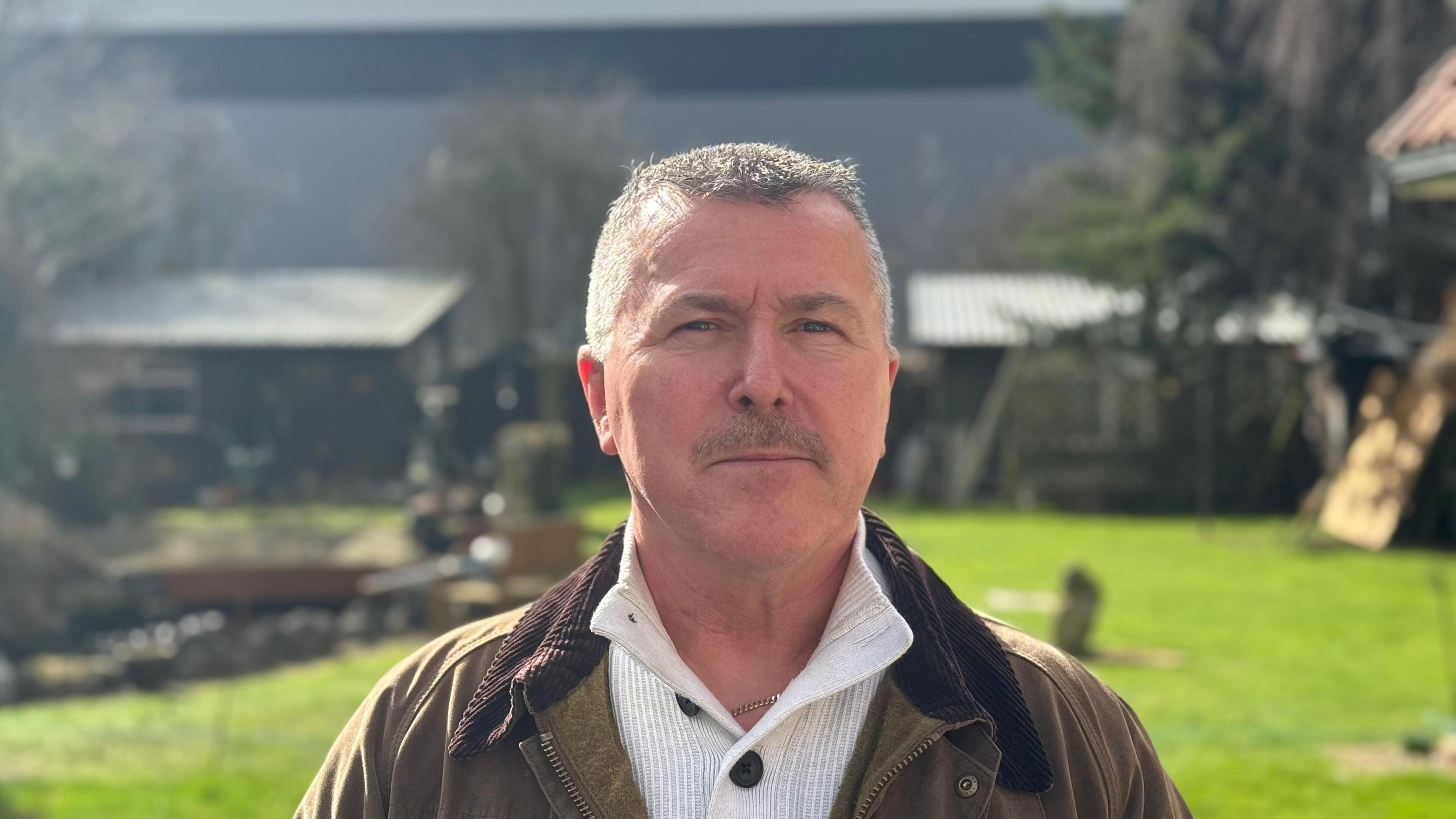 Alan Thomas is wearing a khaki jacket over a white jumper. He is looking directly at the camera. He has short grey hair. He is standing in his back garden and the large warehouse can be seen in the background.