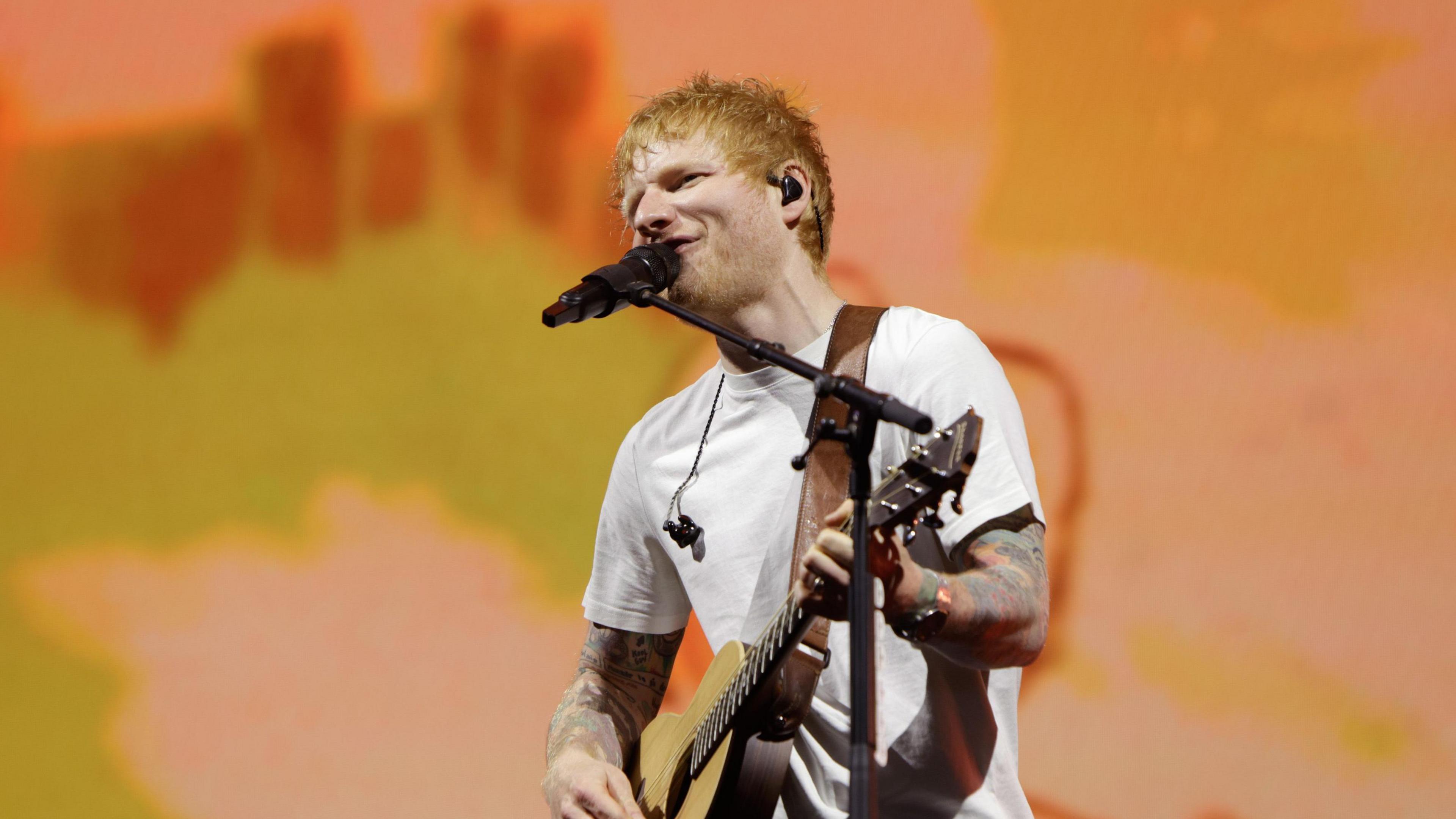 A red-haired Ed Sheeran sings into a microphone while playing a guitar. He is wearing a white tee-shirt