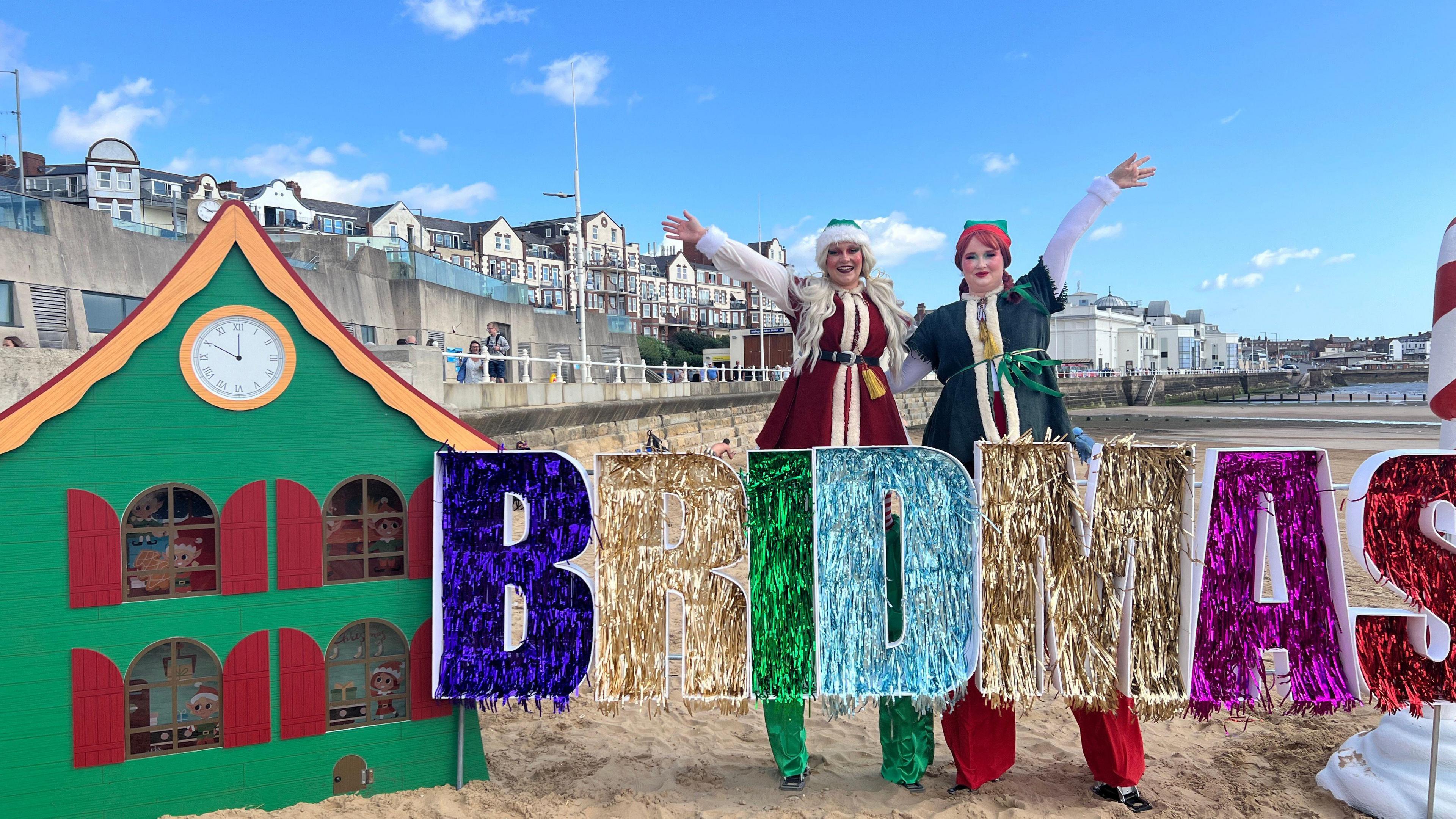 Two women dressed as elves on stilts stand next to a tinsel 'Bridmas' sign