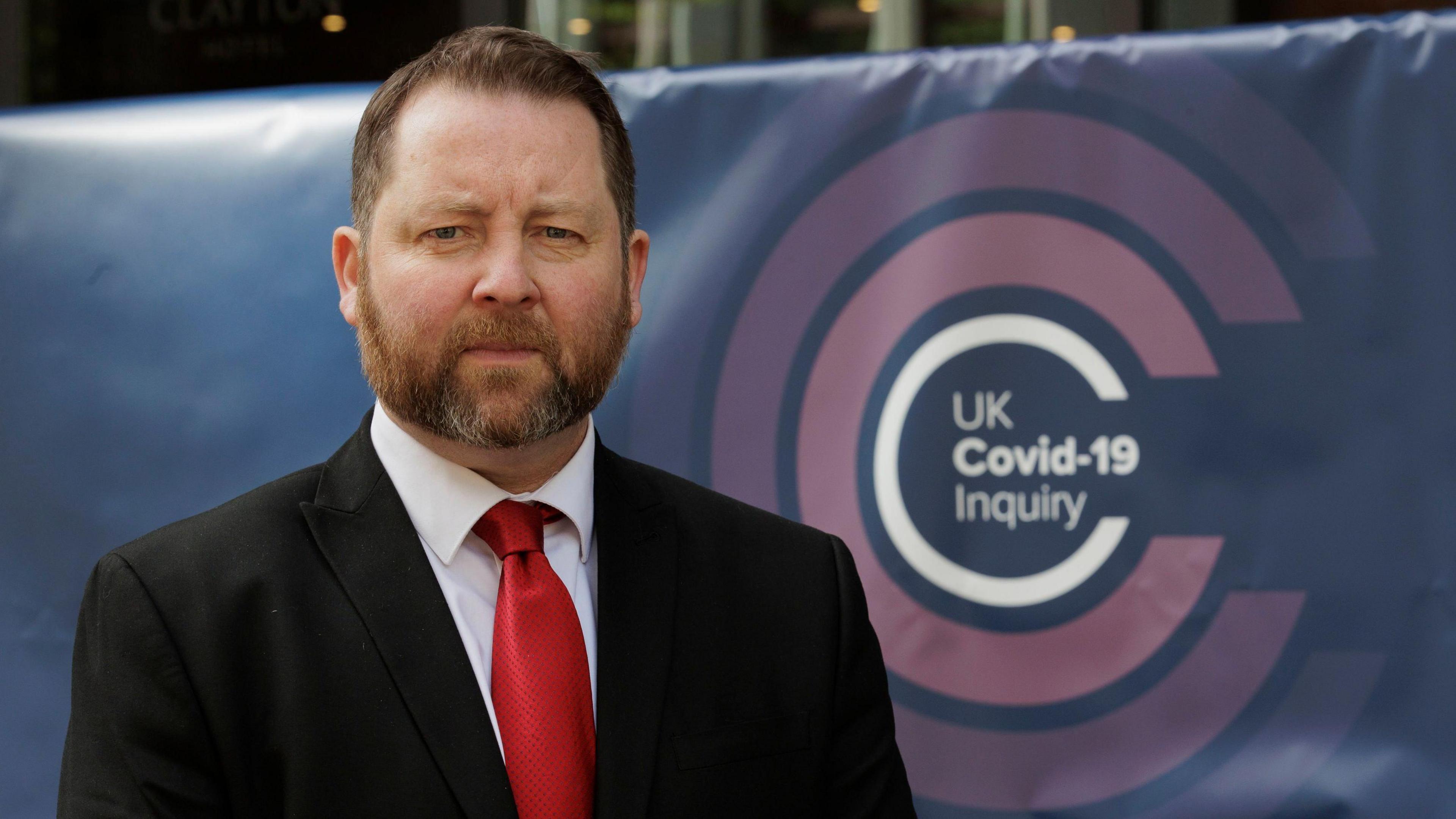 Eddie Lynch has short, reddish brown hair and a greying beard.  He is wearing a black suit, a white shirt and a red tie.  He is standing against a navy blue banner which shows the inquiry's pink and white logo. 