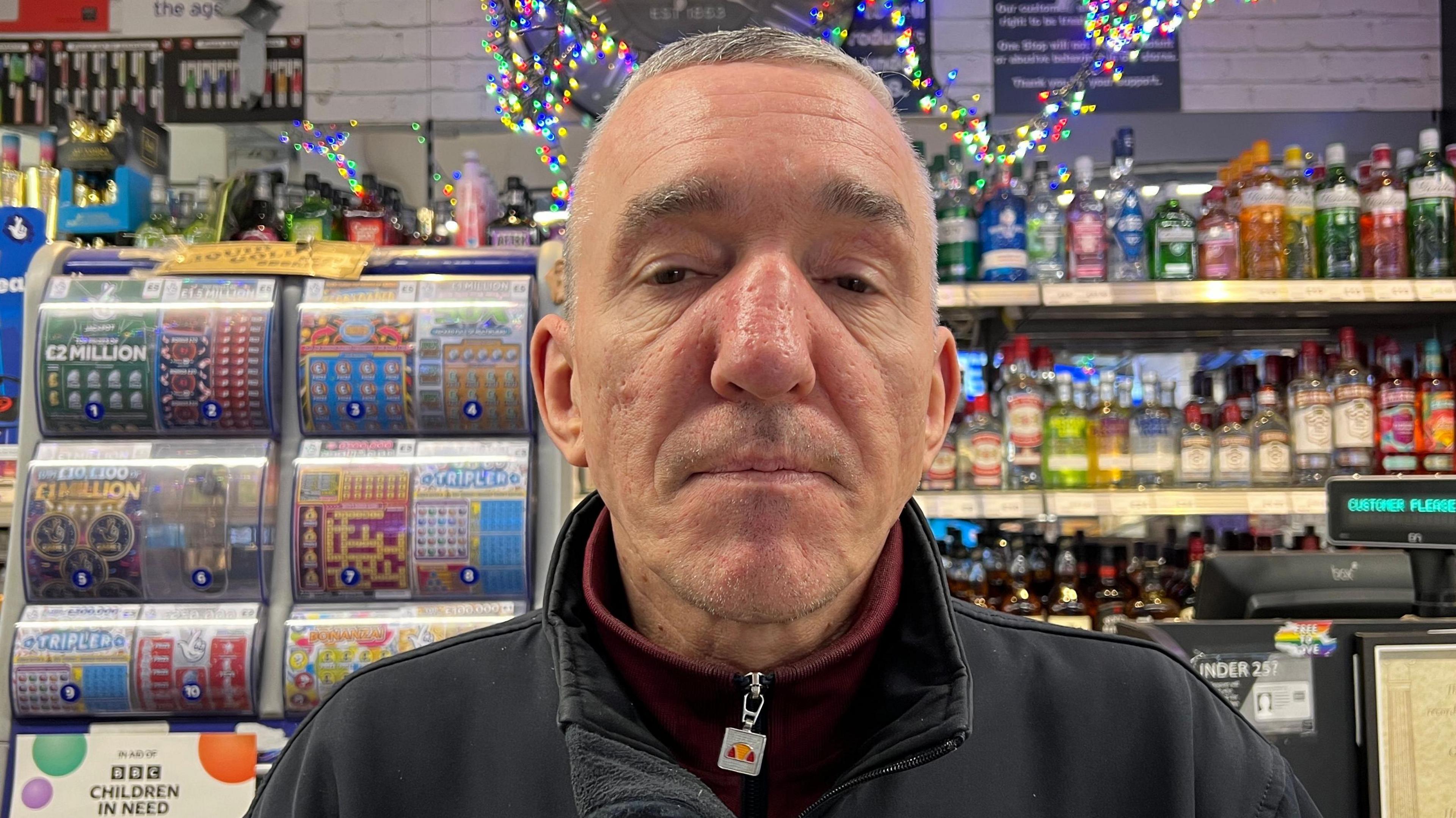 A man standing in front of a kiosk in his shop. In the background, spirits can be seen on shelves and there is a stand full of lottery scratch cards. The man is wearing a black jacket with a red jumper underneath. He has brown eyes and brown hair.
