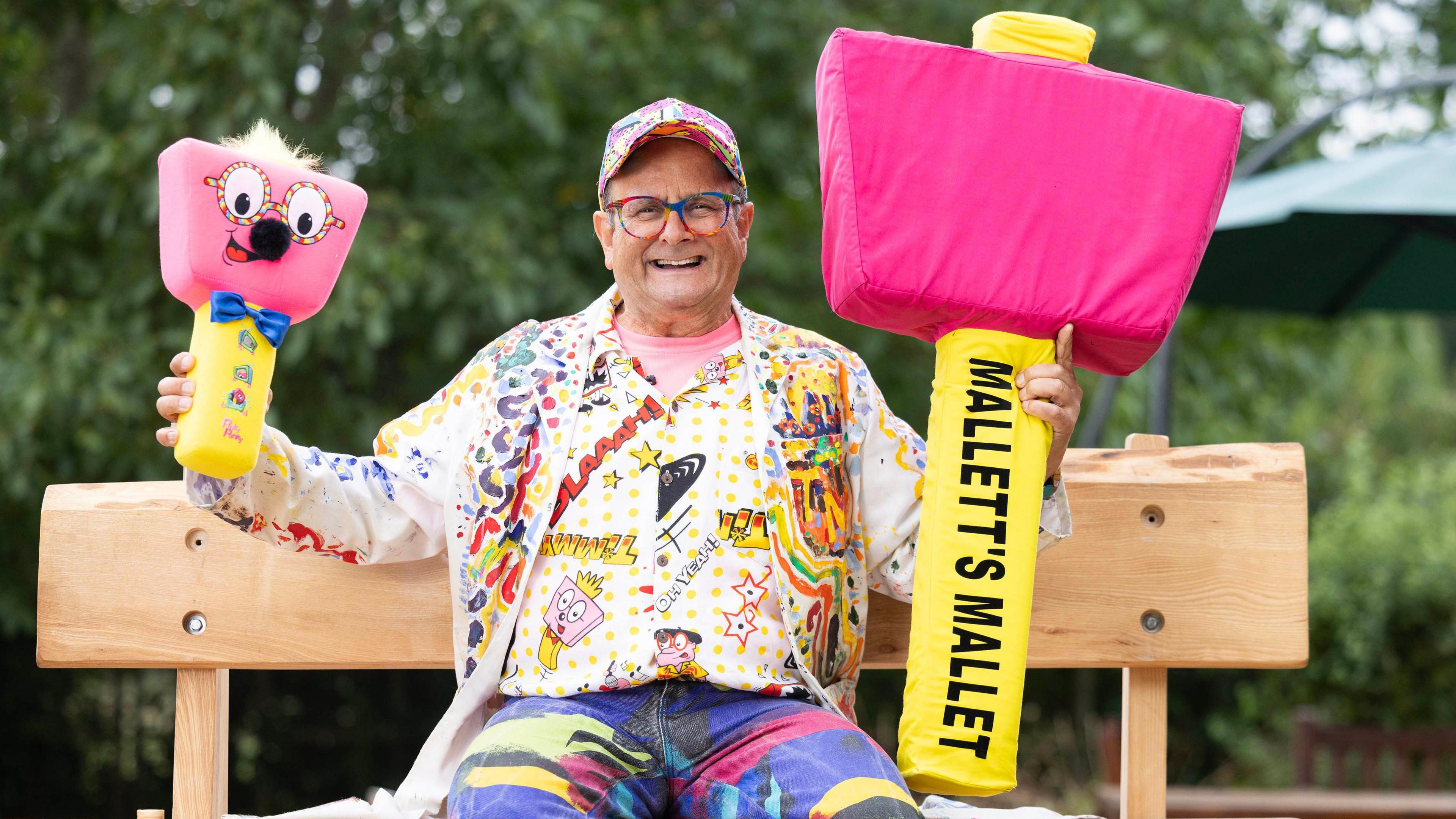 Timmy Mallett in multi-coloured shirt and trousers, sitting on a wooden bench outdoors, holding two pink and yellow toy mallets