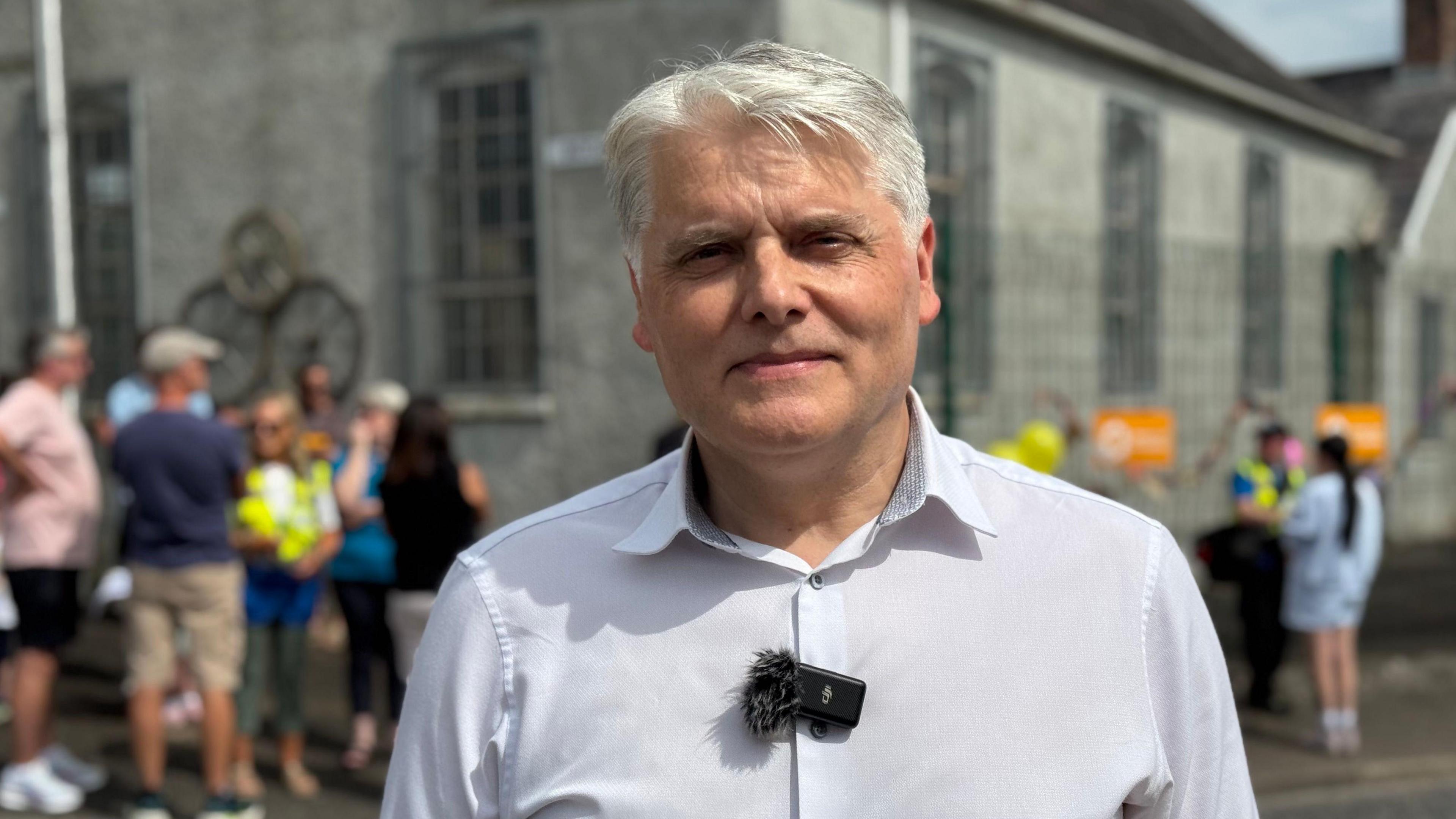 Kieron has neat grey hair, roughly parted. Behind him is a chirch and he is wearing a pale shirt.