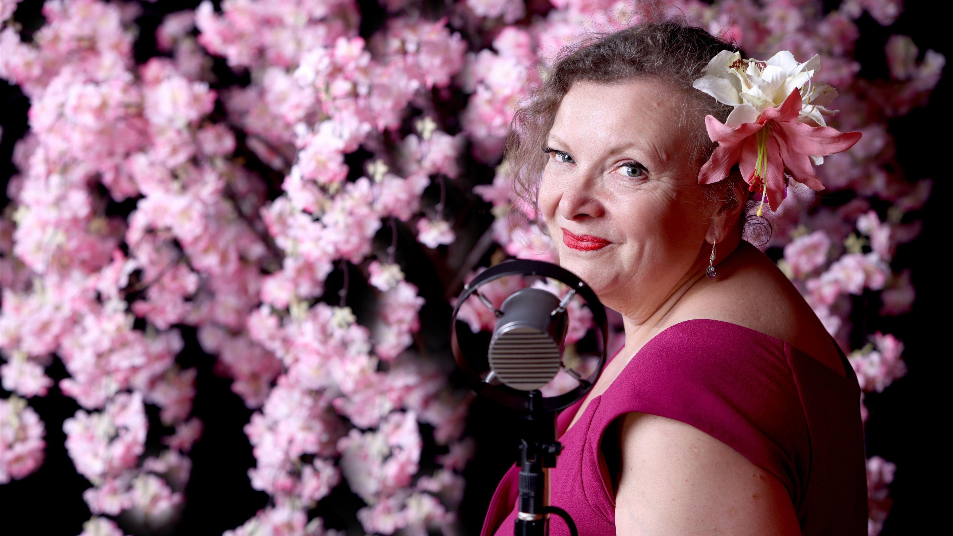 May Blossom, the Vintage Songbird, standing in front of a number of pink flowers. She is looking at the camera and standing in front of a 1940s style microphone, wearing a bright pink dress, with pink flowers in her hair.