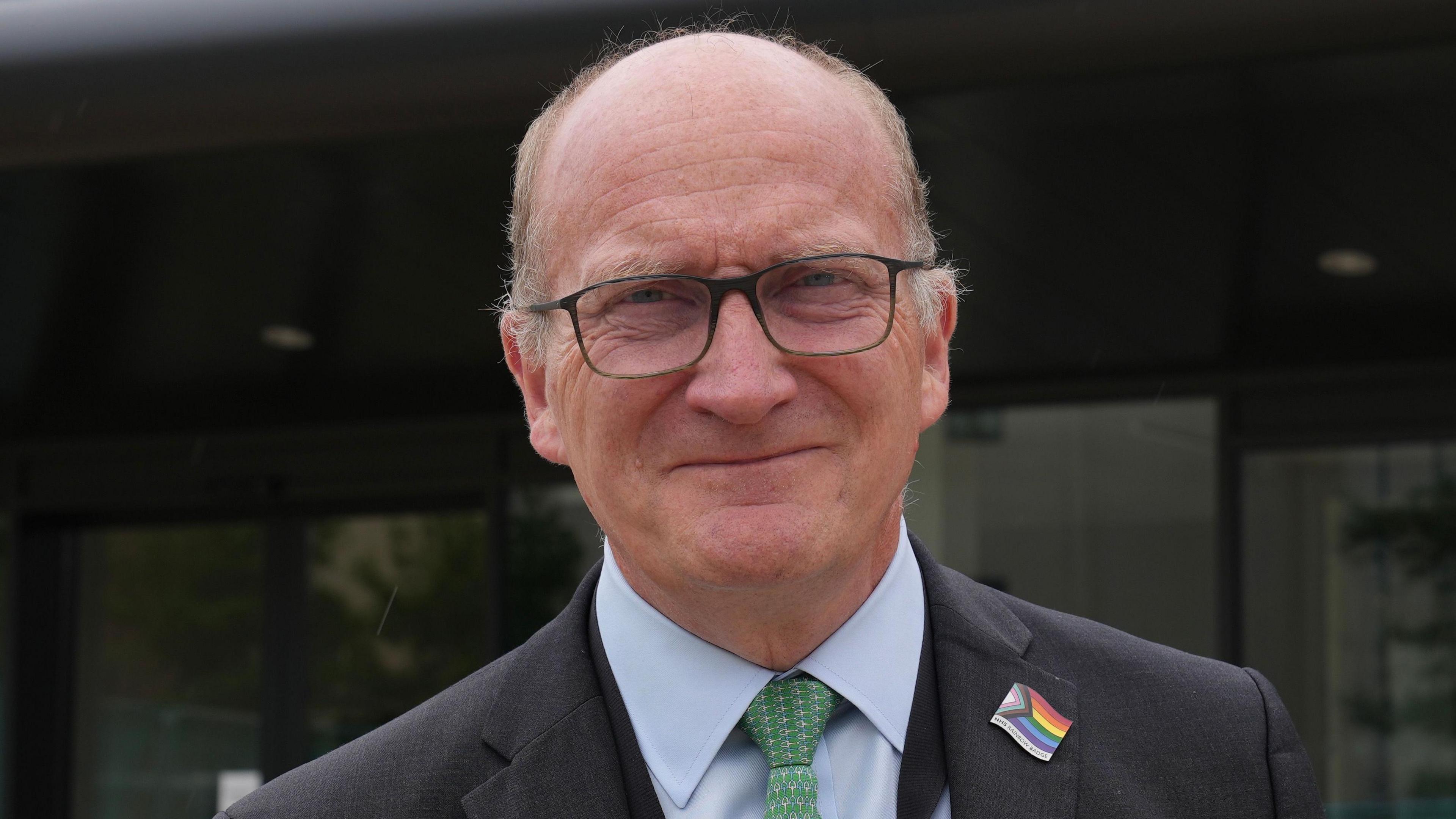 A man wearing a suit and tie smiling at the camera. He is wearing a LGBTQIA+ badge. He is balding and is wearing dark framed glasses, a dark suit, light blue shirt and green tie.