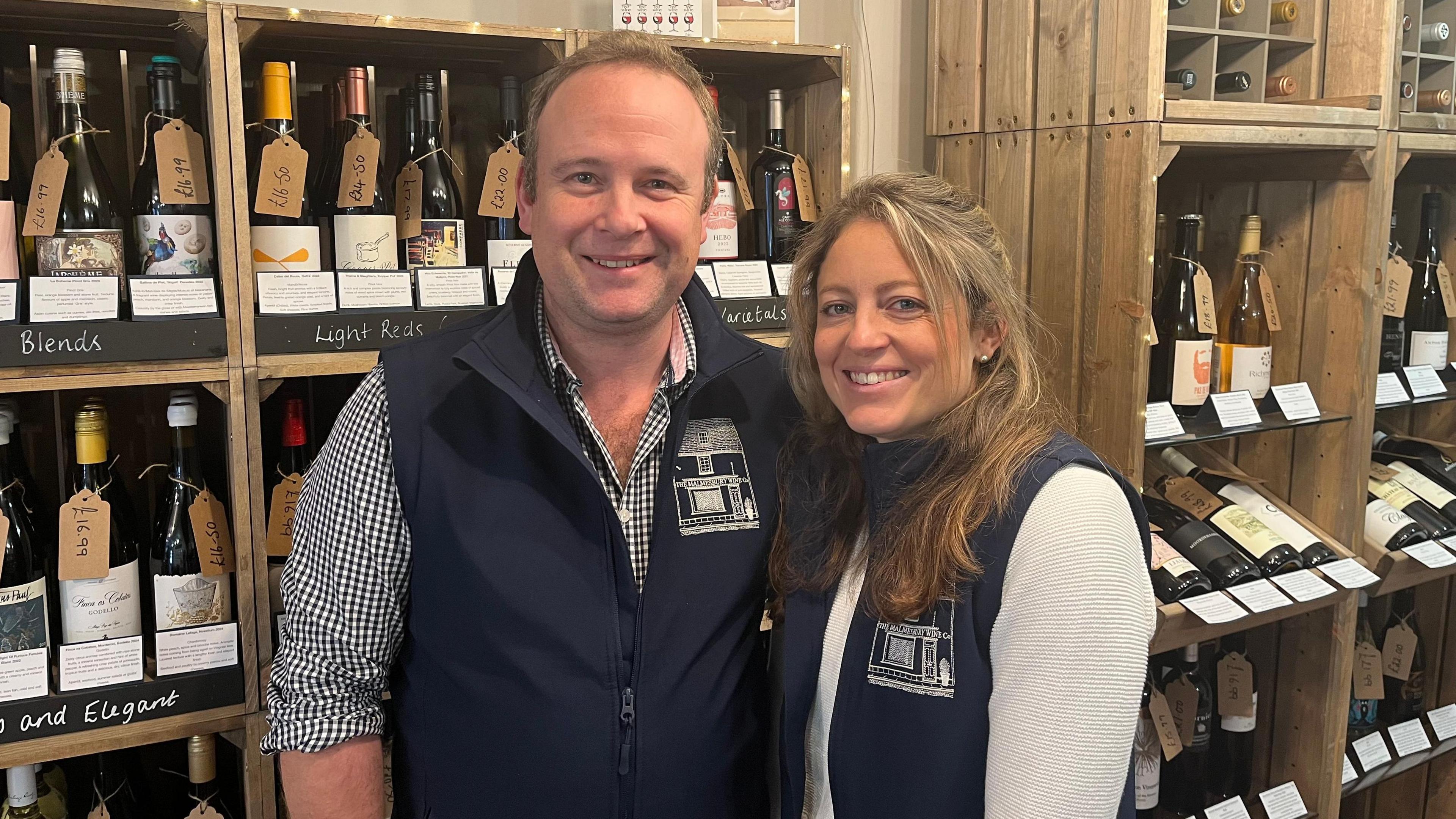 Man and woman both with fair hair and navy blue sleeveless fleeces smiling together with bottles of wine in the background