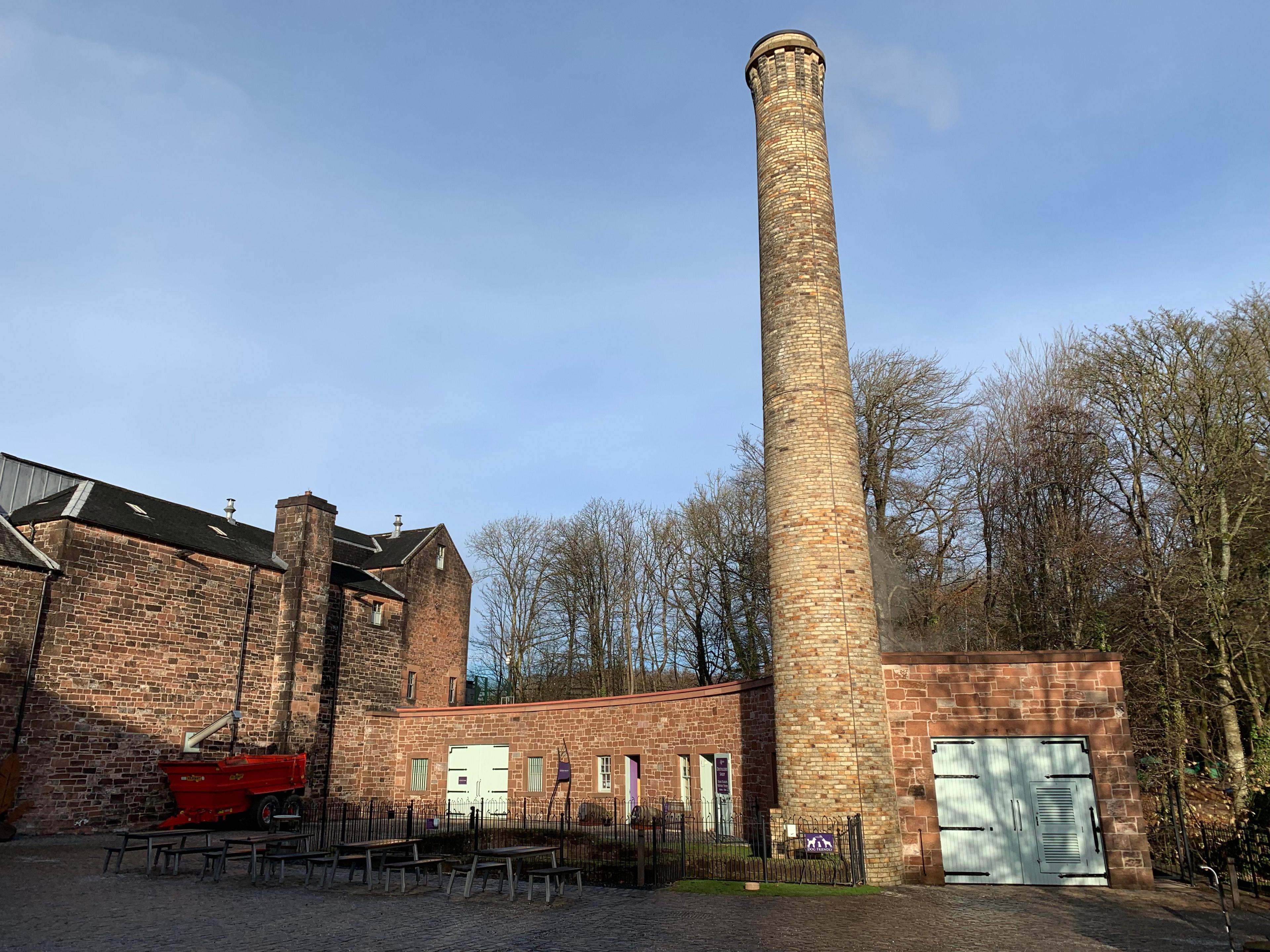 A tall brick tower at a distillery next to two lower sandstone buildings on a lovely blue skied day