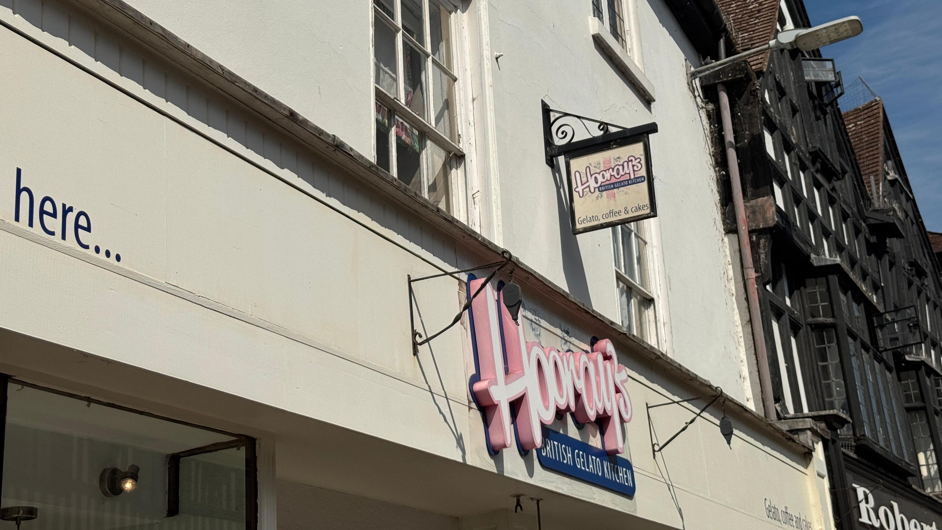 The outside of Hooray's. It is in the historic town of Stratford and the building next to it looks to be of Tudor-style with a black and white facade. Hooray's is a white building with a colourful pink and blue logo.