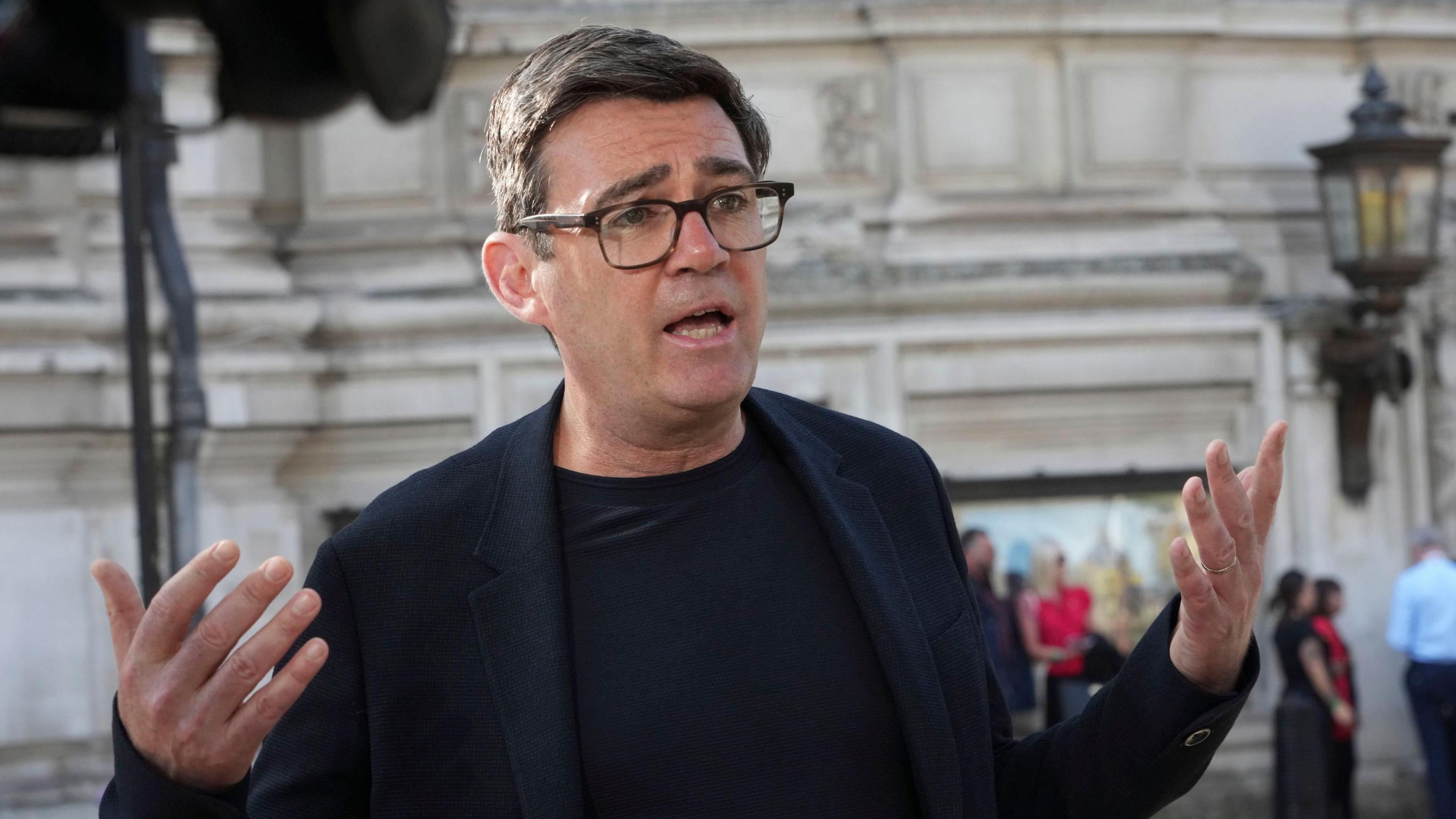 Andy Burnham Mayor of Greater Manchester speaks to the media outside Central Hall in Westminster. He is wearing a black blazer and t-shirt, and black glasses with his hands held aloft.