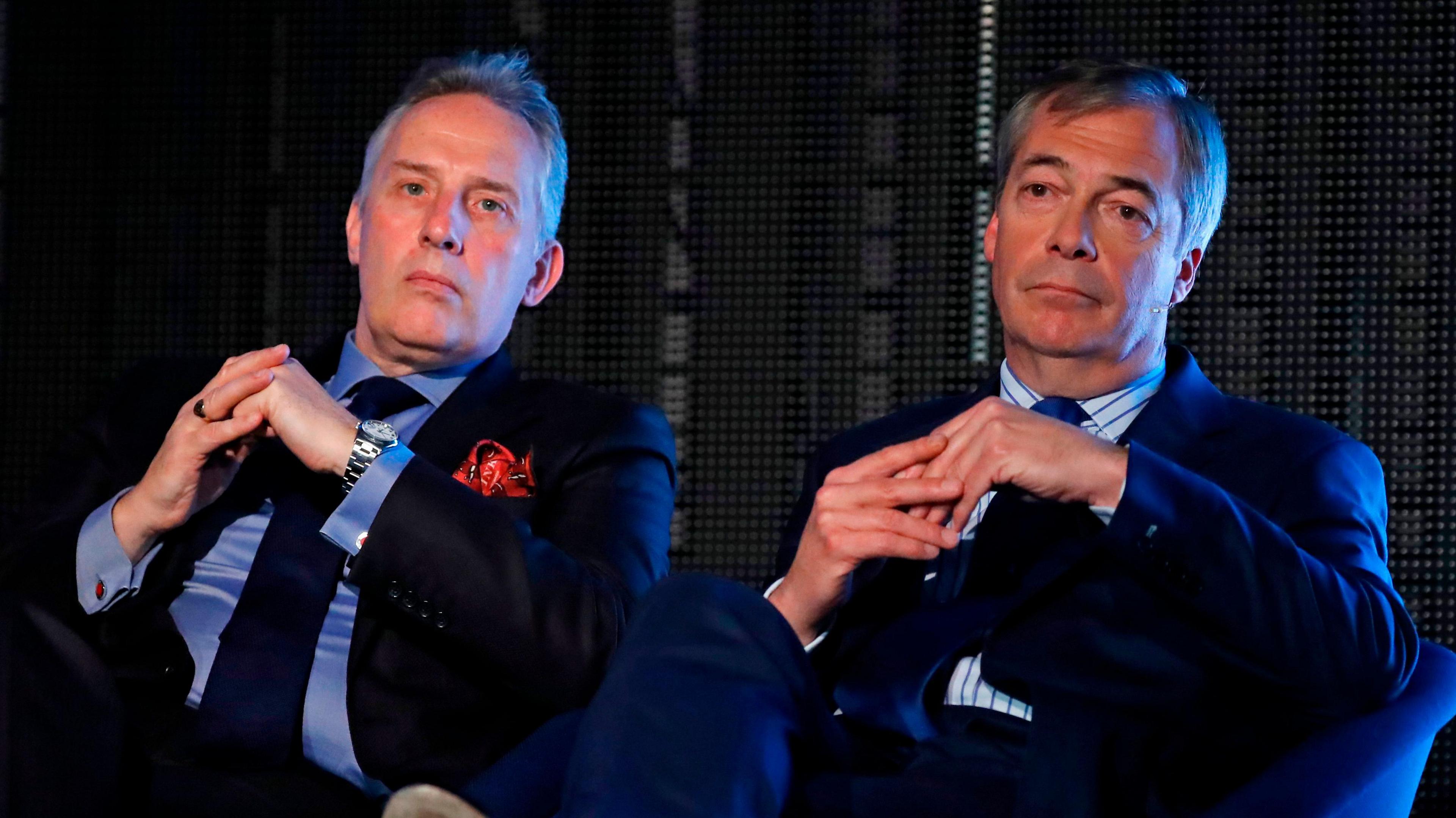 Ian Paisley sits beside Nigel Farage. Both men are wearing suits, clasping their hands and looking beyond the camera.