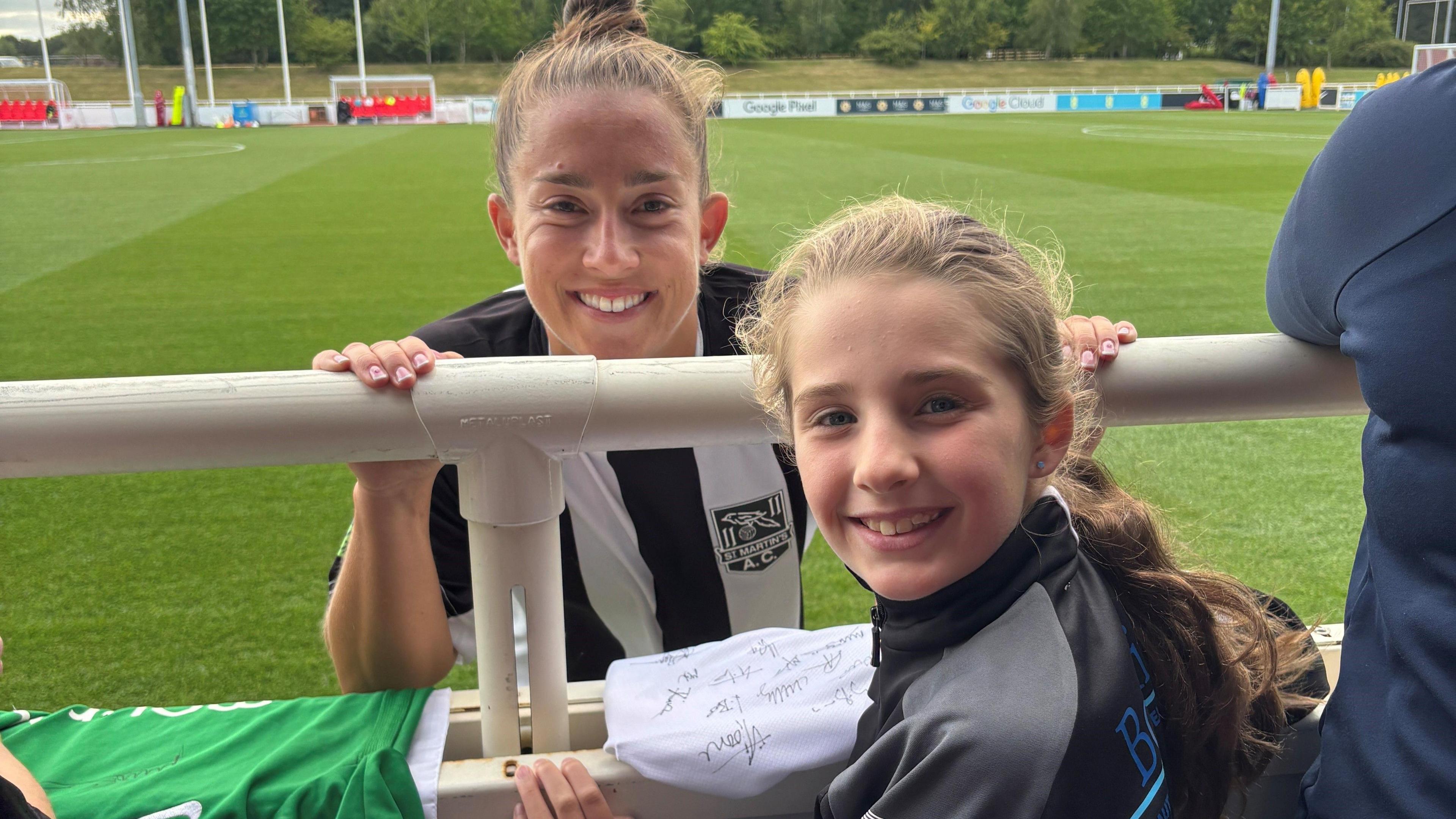 A smiling young girl with a long ponytail poses with female footballer Maya Le Tissier. The player is wearing a black and white striped football shirt.  