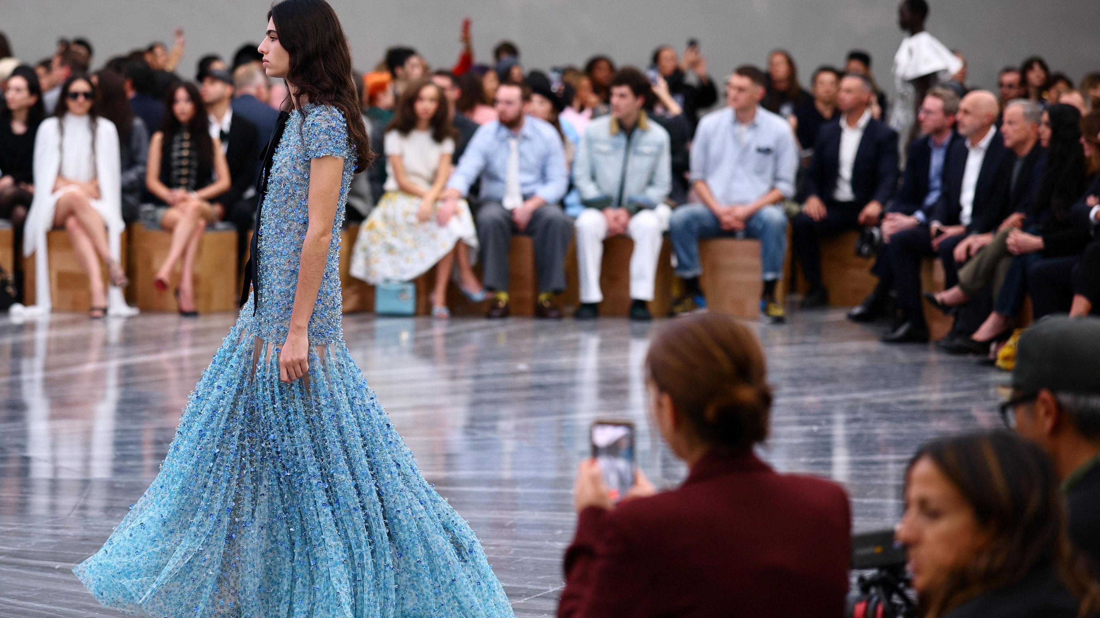 A woman in a blue dress with long brown hair. She is on a catwalk, with the audience behind her.