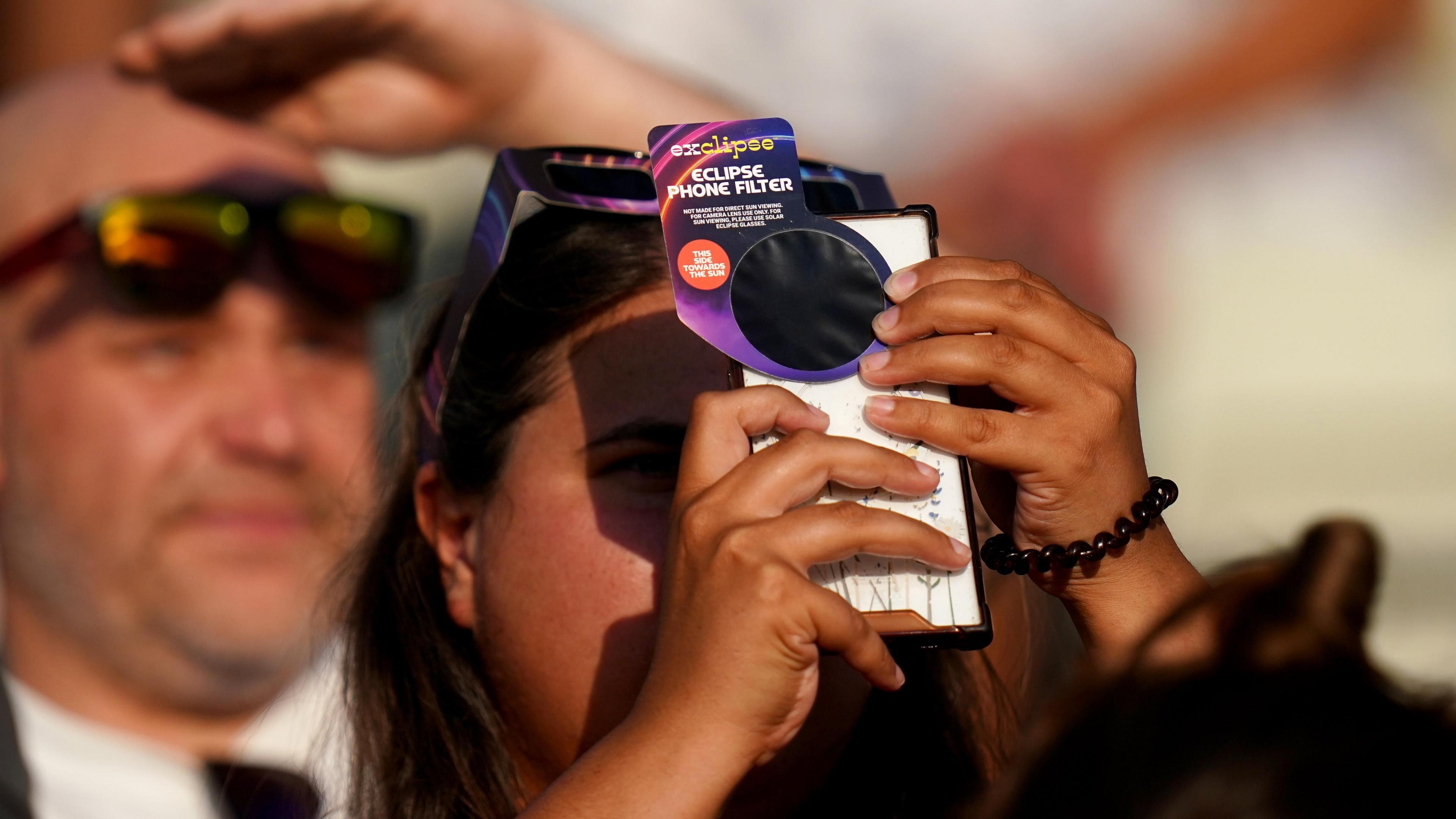 Person holding a mobile phone fitted with an eclipse phone filter in the foreground, aiming it upwards. Another person nearby wears eclipse-viewing glasses and shades their eyes with a raised hand. The scene is outdoors in bright sunlight, with a crowd visible in the background.
