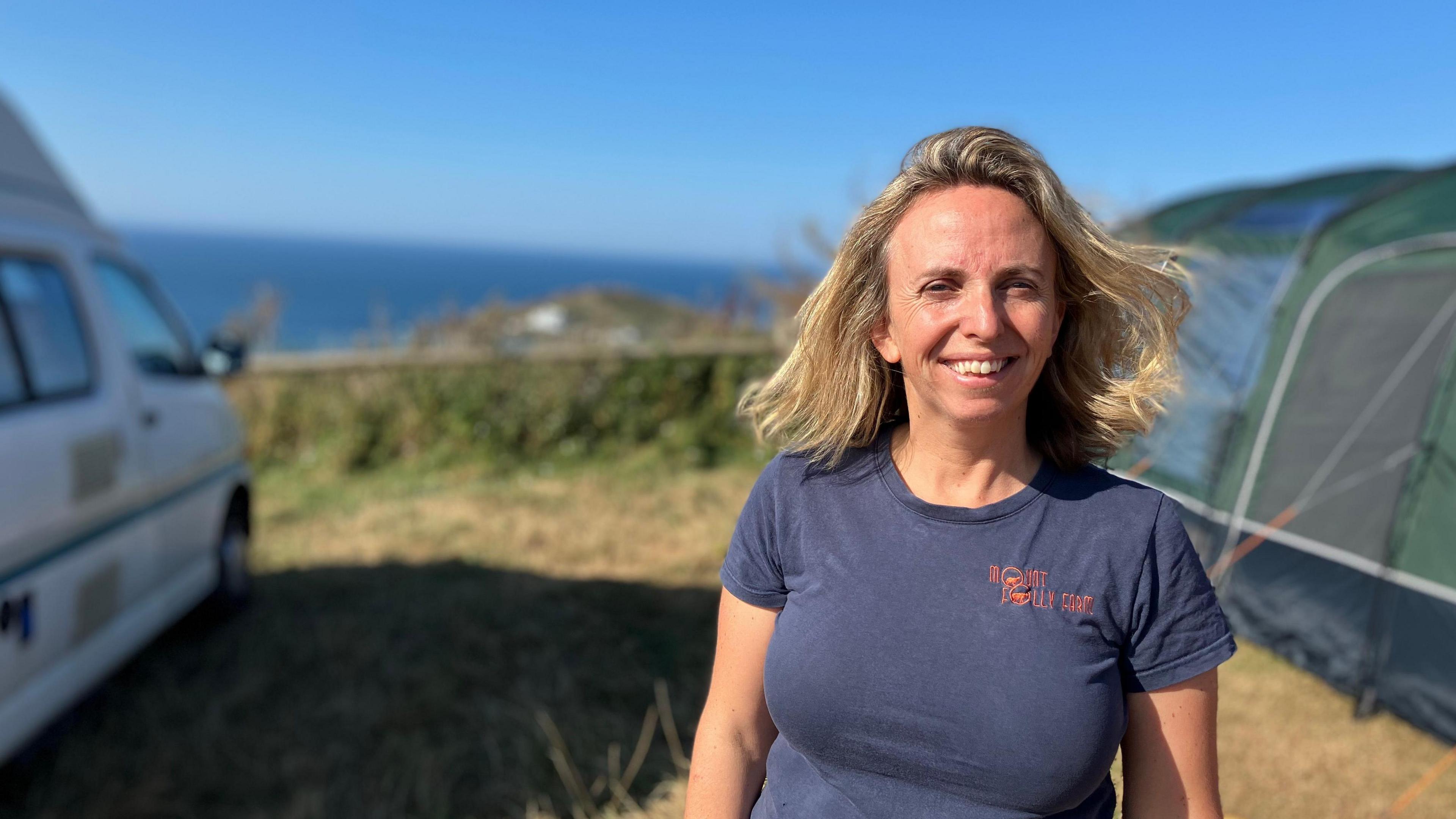 A woman stands in a field overlooking the coast, she is in between a tent and a campervan.