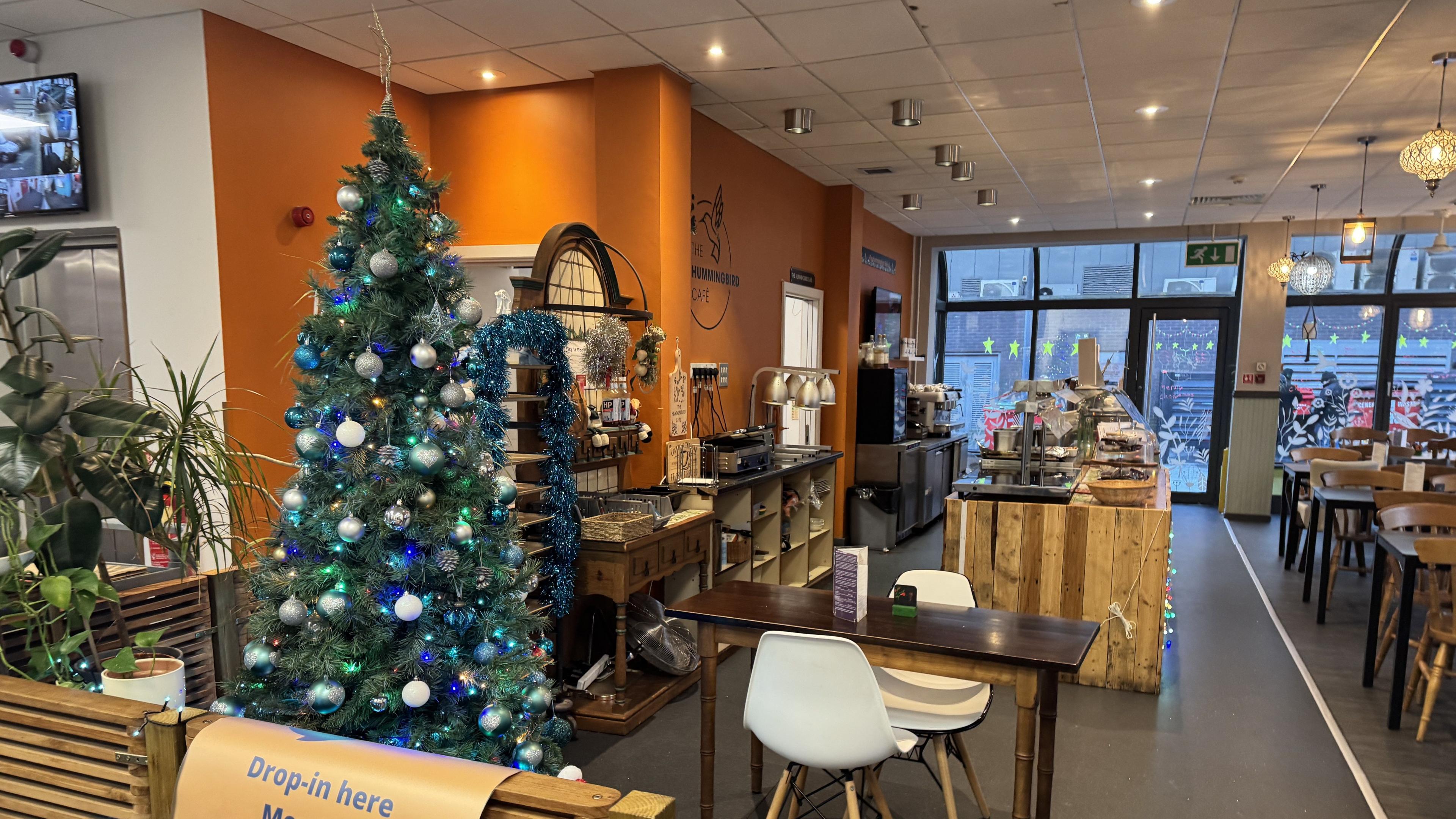 A cafe with orange walls. there are wooden tables with chairs and a wooden counter. There is a green Christmas tree with blue and silver baubles