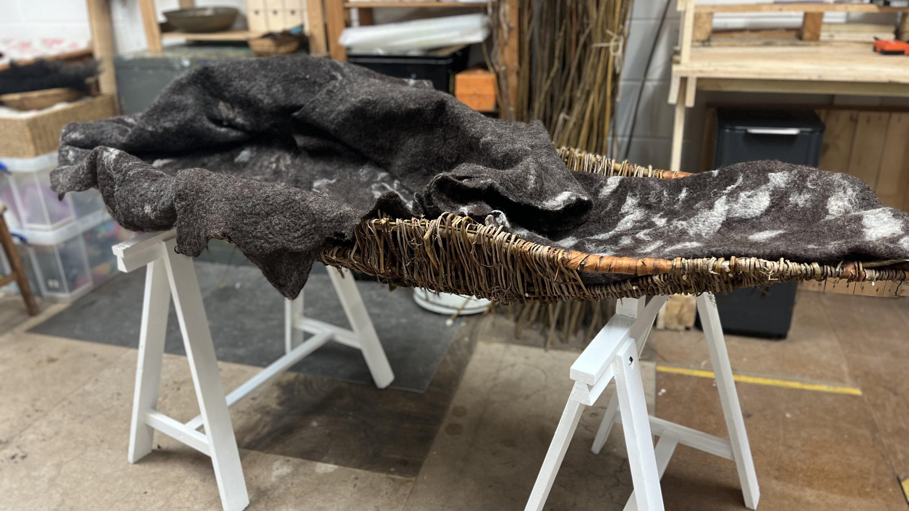 A woollen, black-and-white burial shroud sits in a wicker basket in an artist workshop. It is displayed on top of a white wooden stand. 