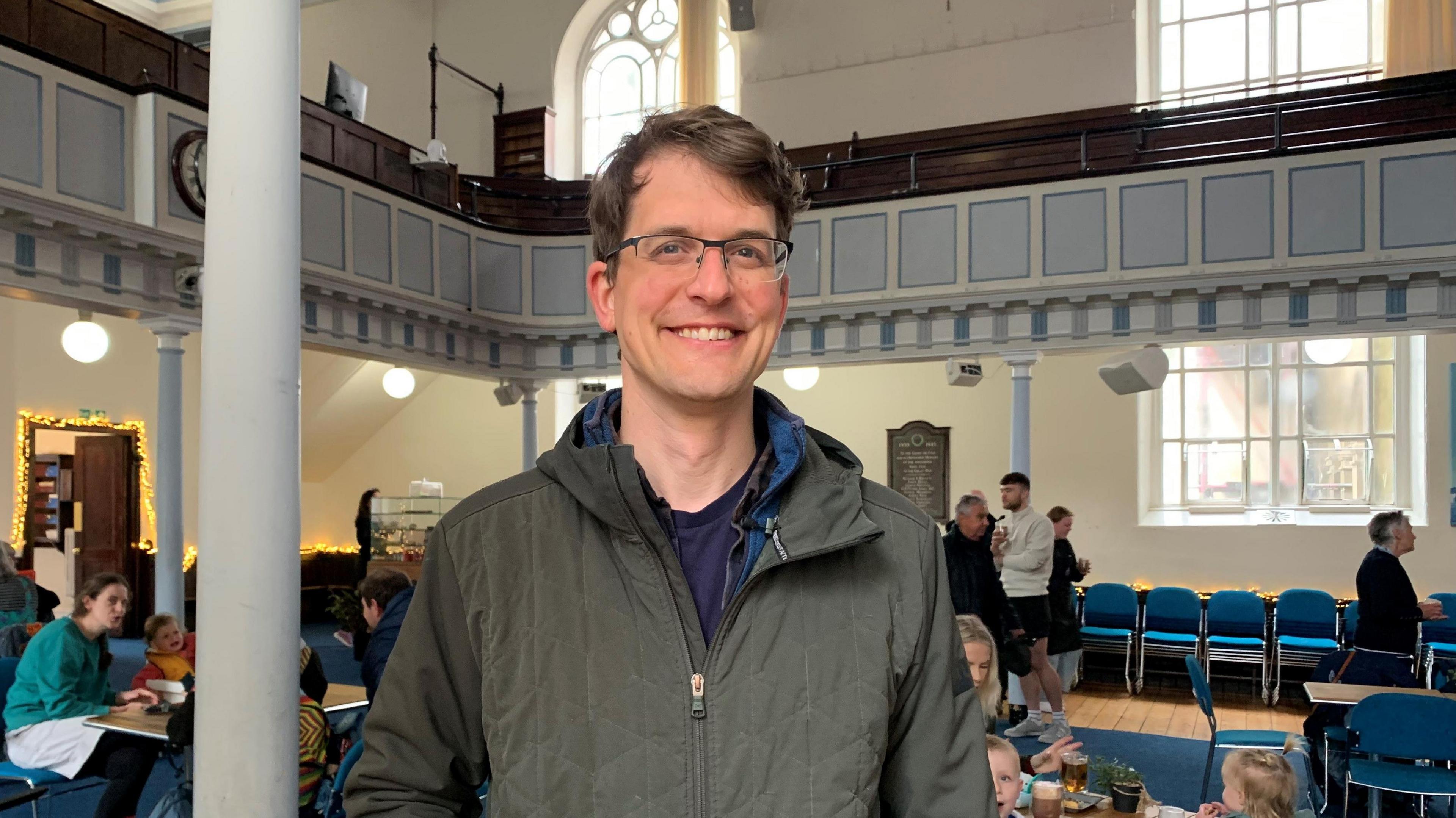 Tom Martin is smiling at the camera on the main floor of the church. There are people sitting behind him and you can see the upper tier of pews and arched glass windows. He is wearing a green jacket, has medium length parted brown hair and has glassed.