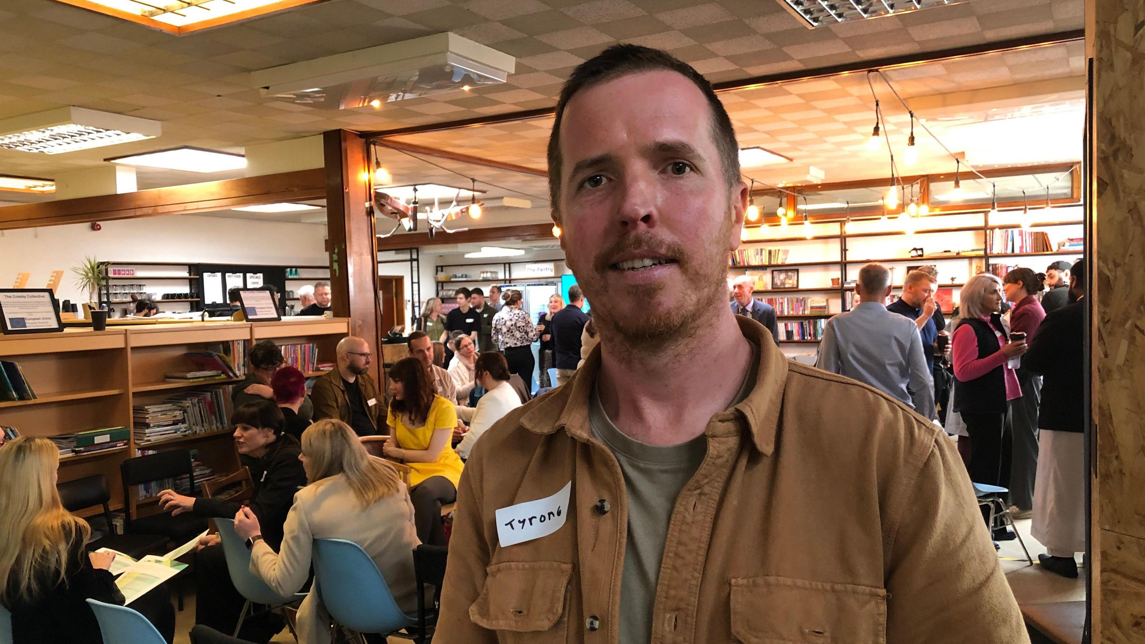 Tyrone Curran smiles as he stands in a well-lit room during a busy networking event. He has short dark hair and a short brown beard. He wears a brown denim shirt over a pale green T-shirt. Groups of people can be seen talking in the background.