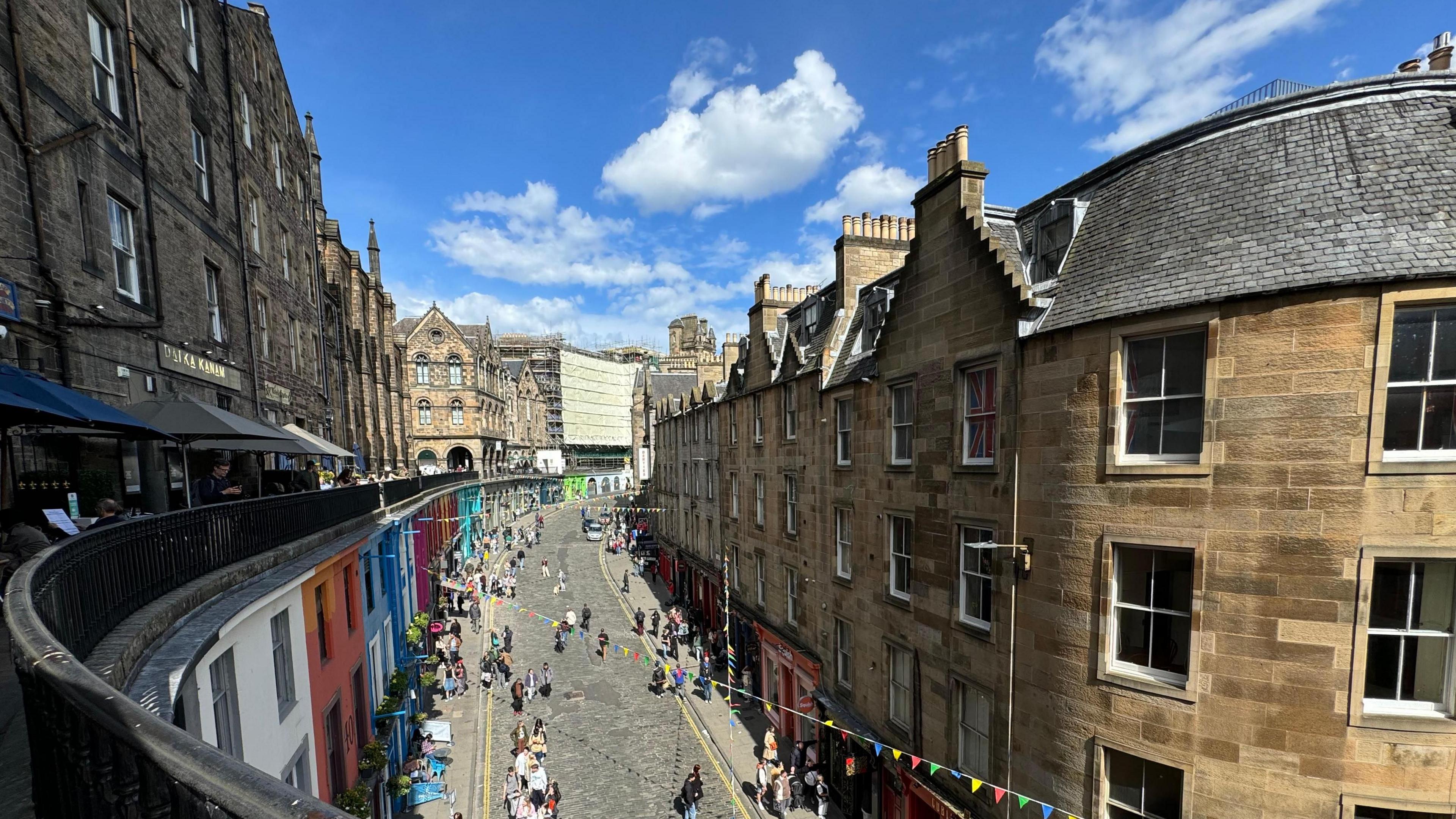 View looking down onto a cobbled pedestrianised street in Edinburgh. Stone buildings are on either side, colourful bunting crosses from one side to another, and sunny skies are overhead